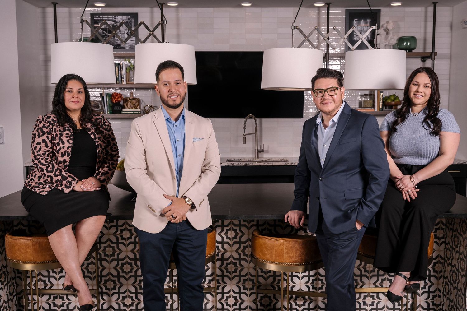 Professional business team members pose together in a modern office setting with patterned tile floor and kitchen area.