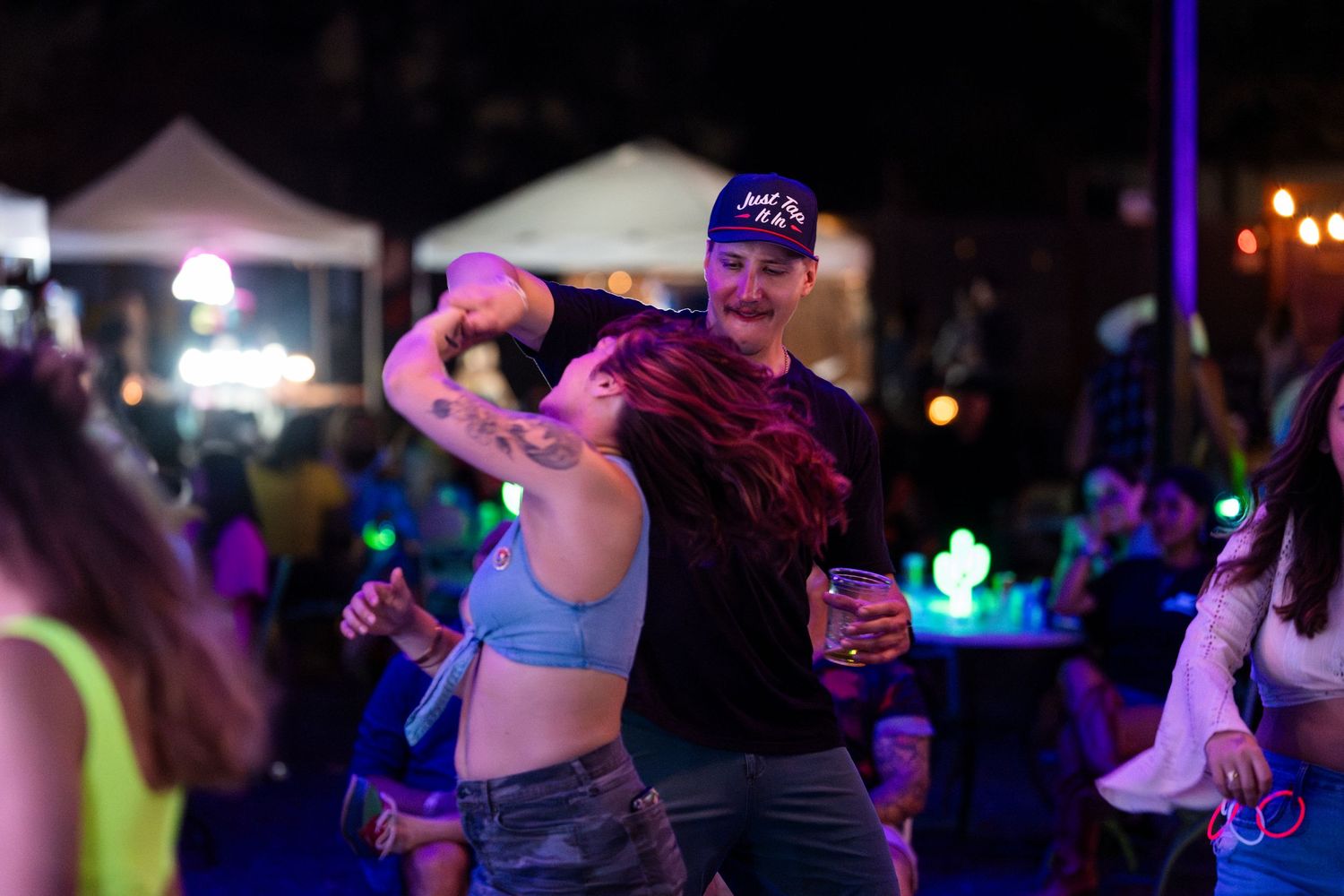 People dancing and having fun at a nighttime outdoor festival with colorful lighting and tents in the background.