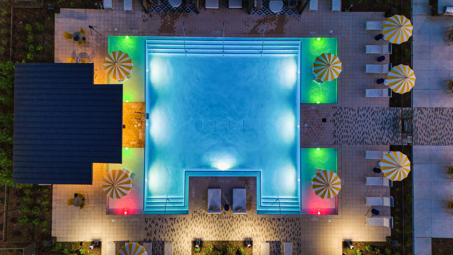 Aerial view of a square swimming pool with colorful lighting and palm-shaped decorative elements at night.