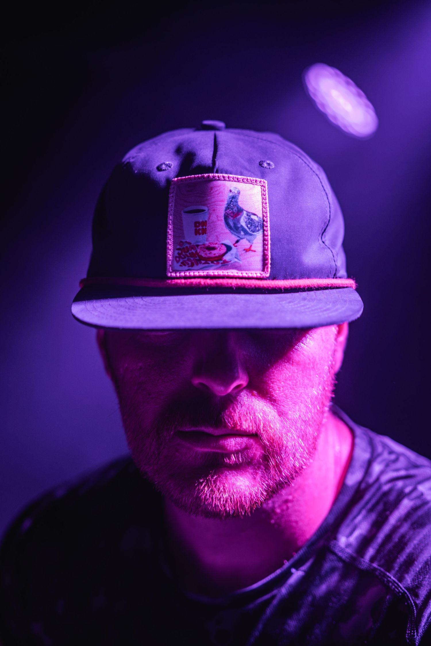 Close-up portrait of person wearing baseball cap under purple stage lighting.