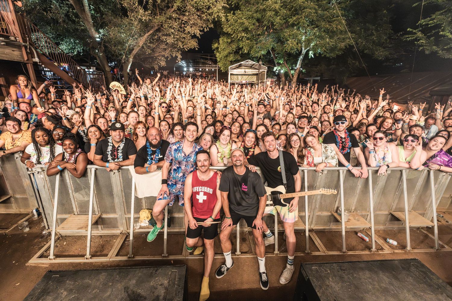 Large excited crowd at an outdoor concert venue with barricades and trees in the background.