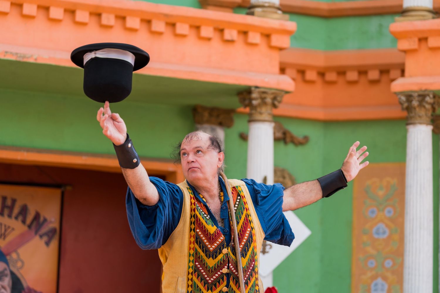 Performer in colorful vest raises black top hat during outdoor renaissance festival show.