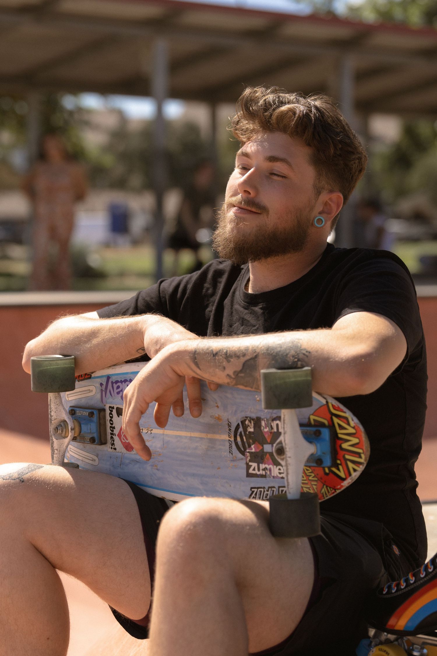 Person holding skateboard at skate park wearing black t-shirt on sunny day.