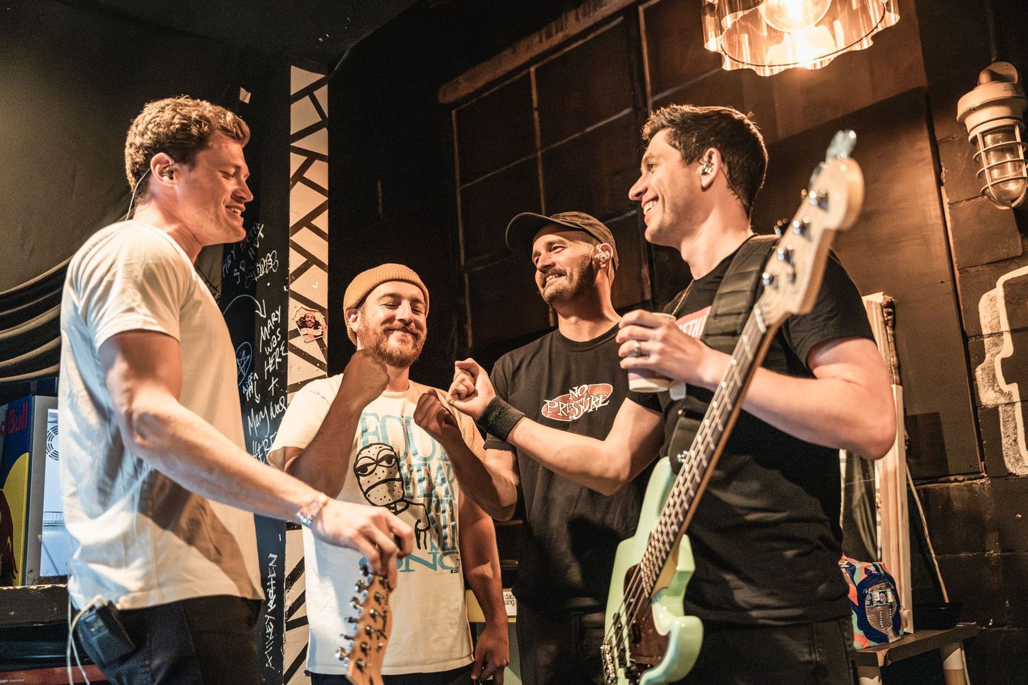 Four band members share a lighthearted moment while performing on stage with guitars and microphones.