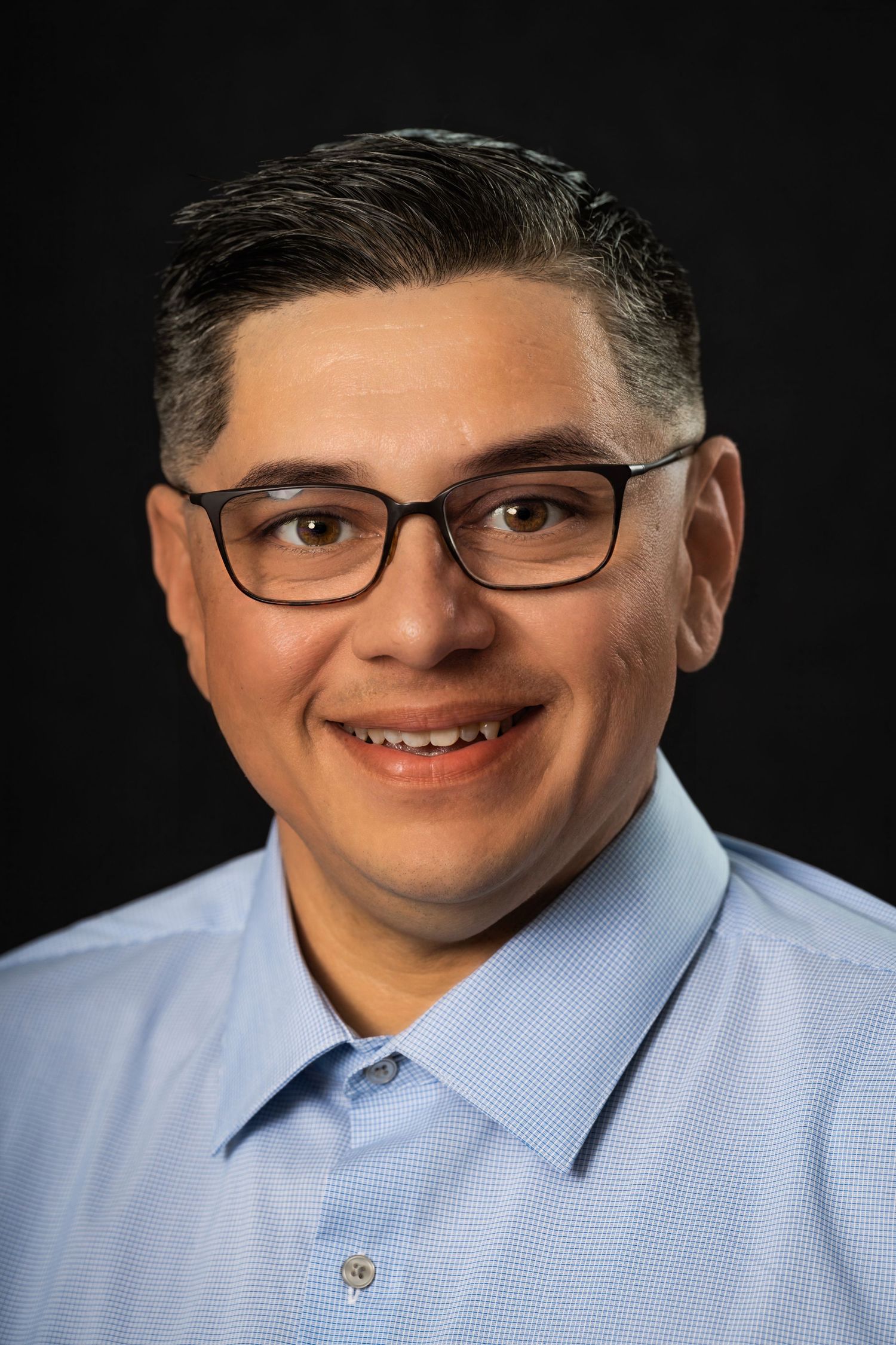 Professional headshot of person in light blue shirt and glasses against dark background.