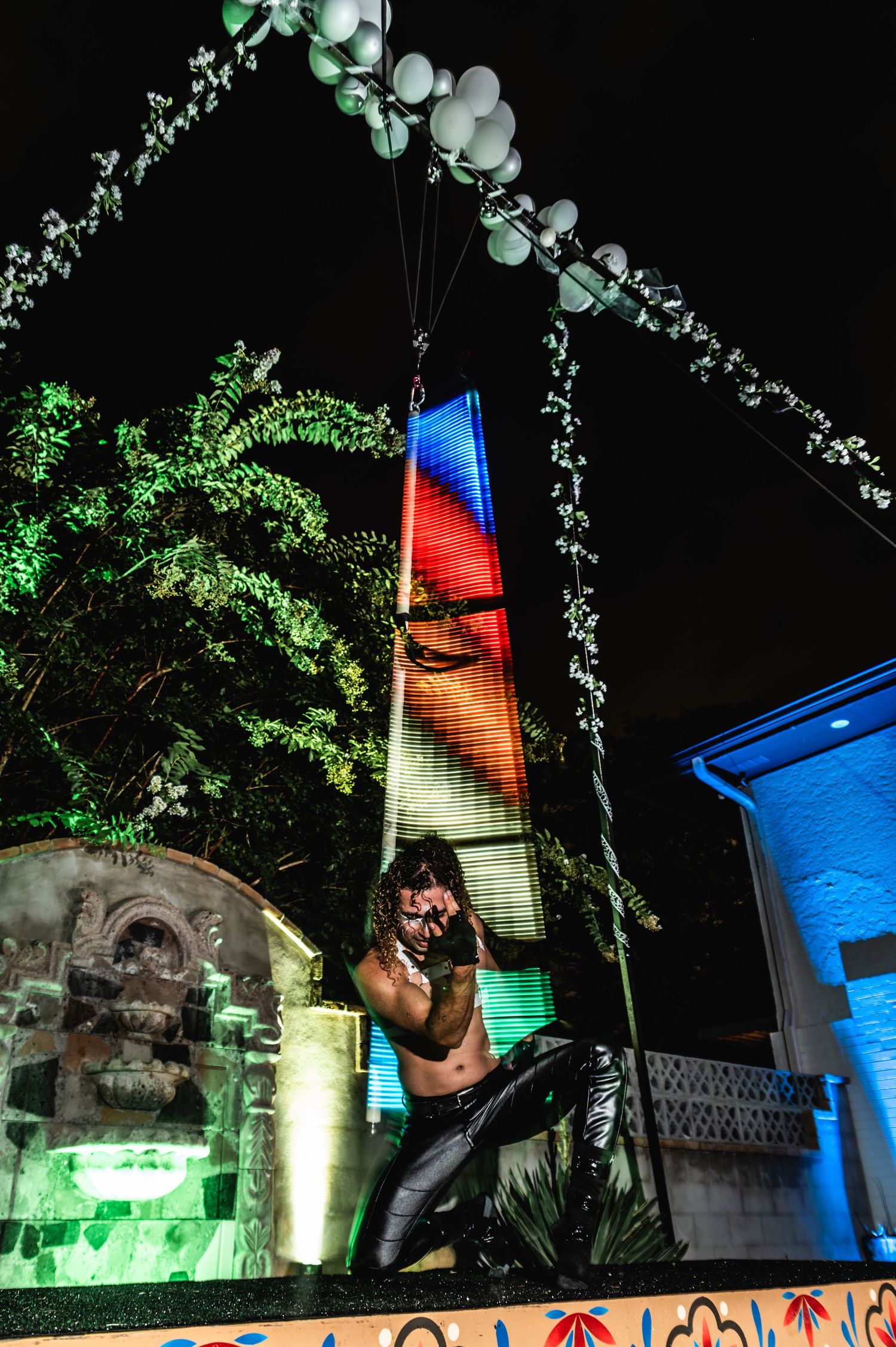 Performer poses on colorfully lit tower structure during nighttime outdoor show.