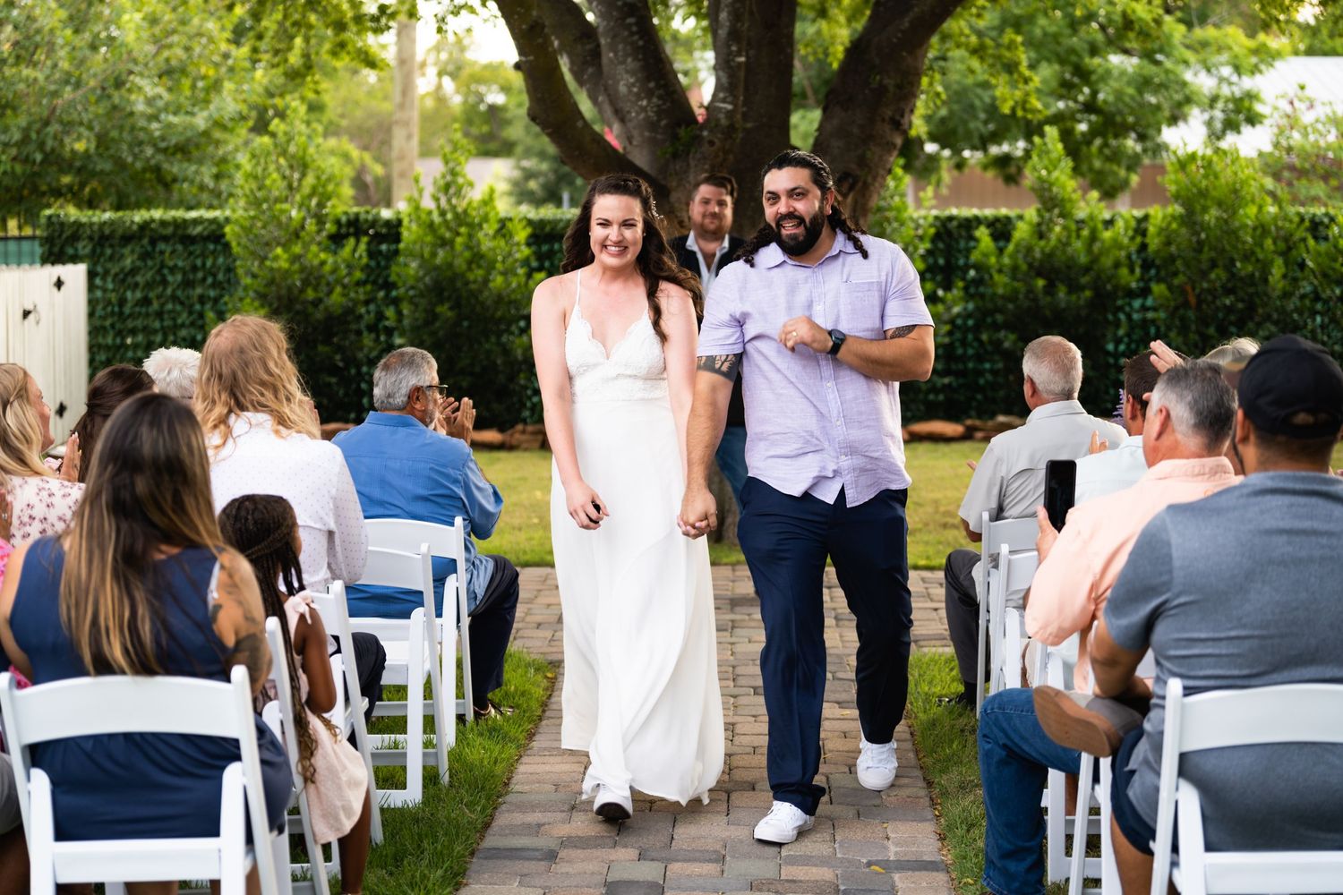 Couple walks down outdoor aisle between white chairs during garden ceremony.