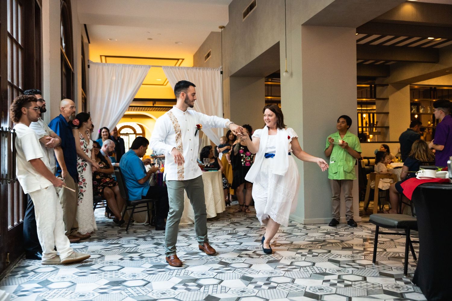 People dance and celebrate in an elegant venue with patterned floor tiles and white curtains.