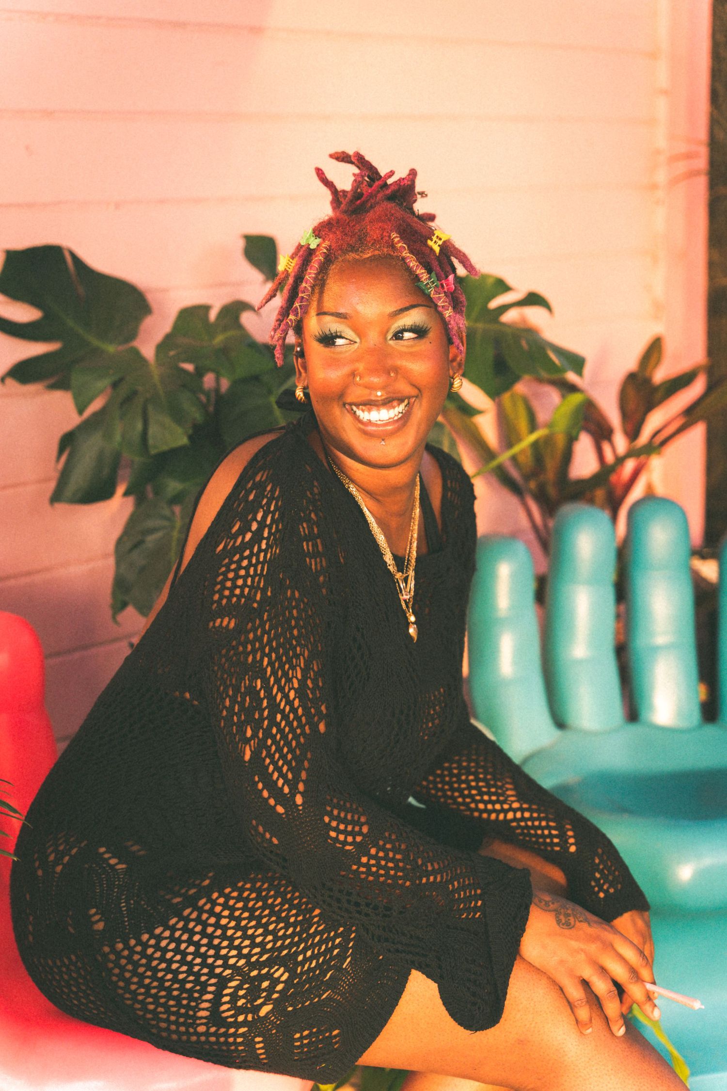 A joyful expression in a black mesh dress and colorful braids sitting next to a large teal decorative hand sculpture.