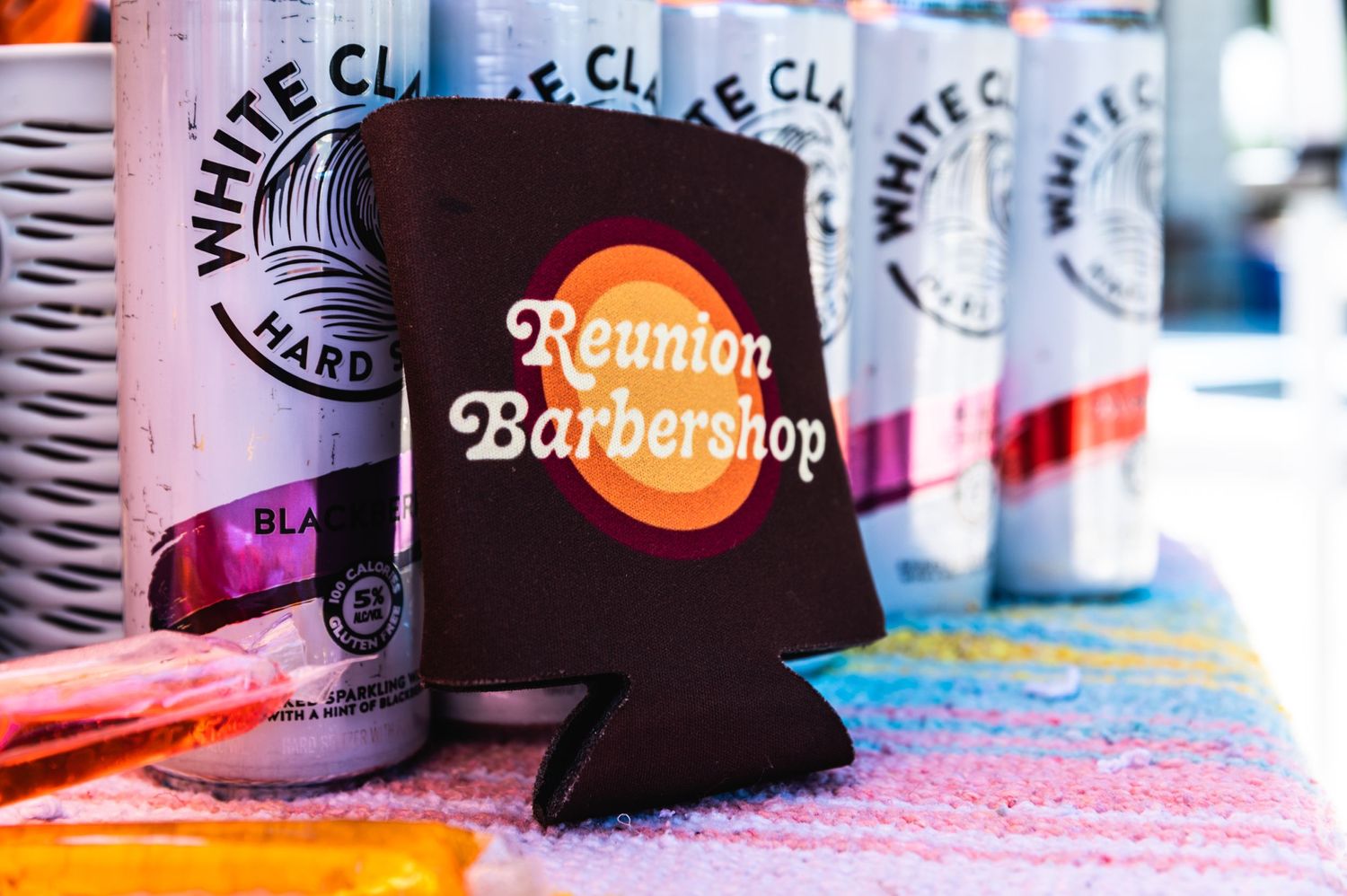 A black drink koozie with 'Reunion Barbershop' logo next to White Claw cans.