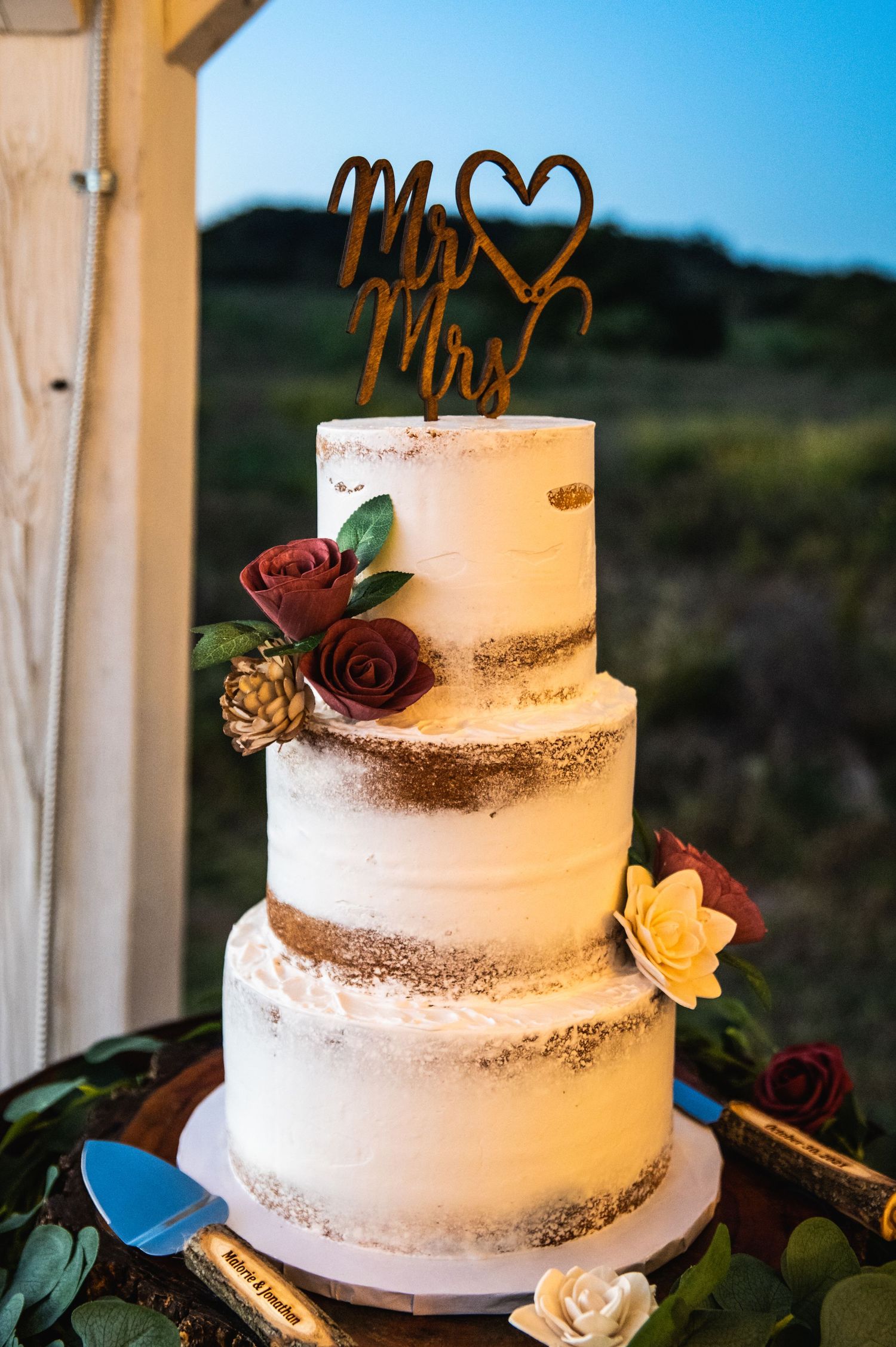 Three-tiered white wedding cake with gold accents, burgundy roses and Mr & Mrs topper against blue sky.