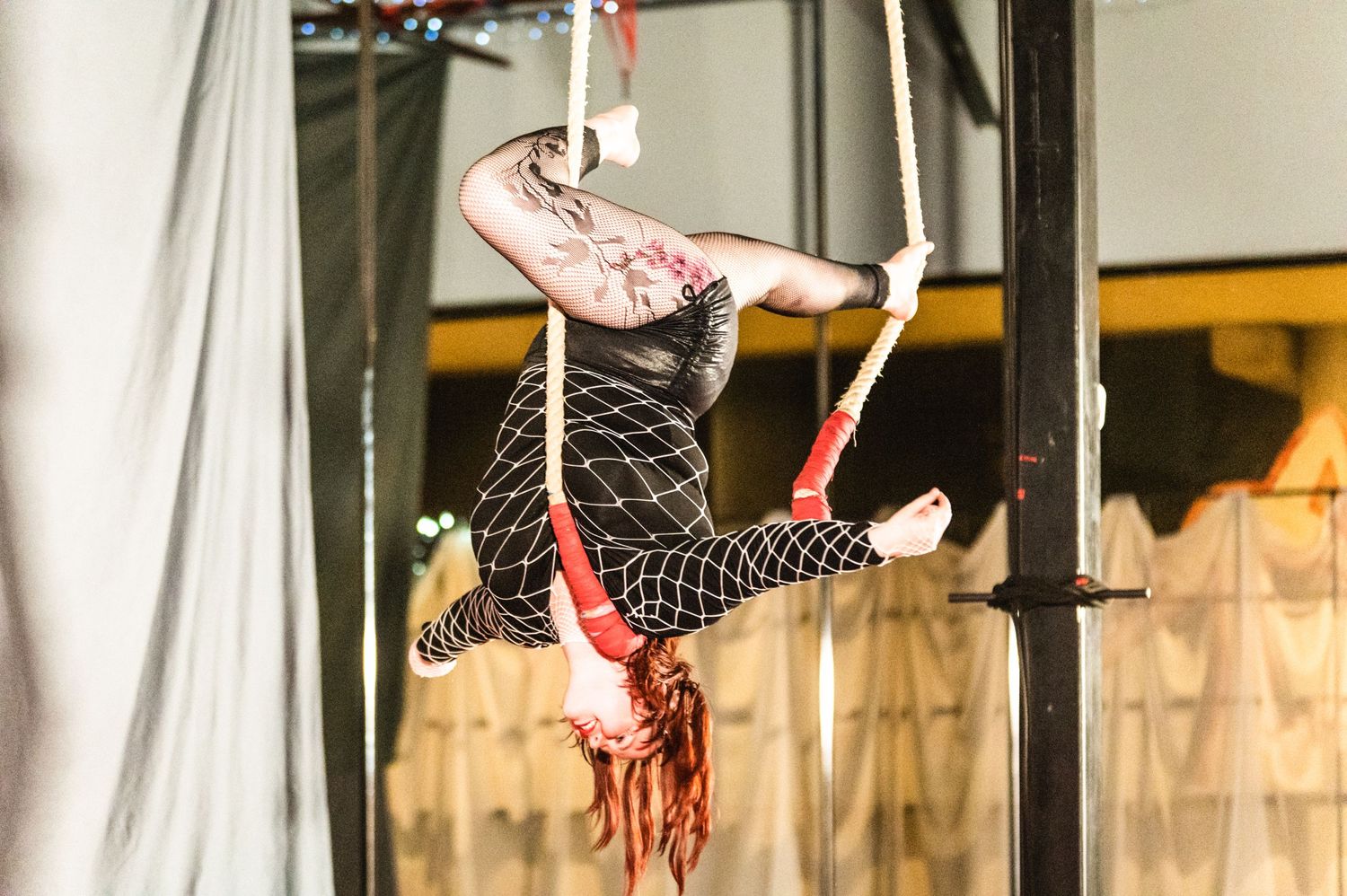 Aerial performer in black and red costume executes an inverted pose on suspended silks during a circus performance.