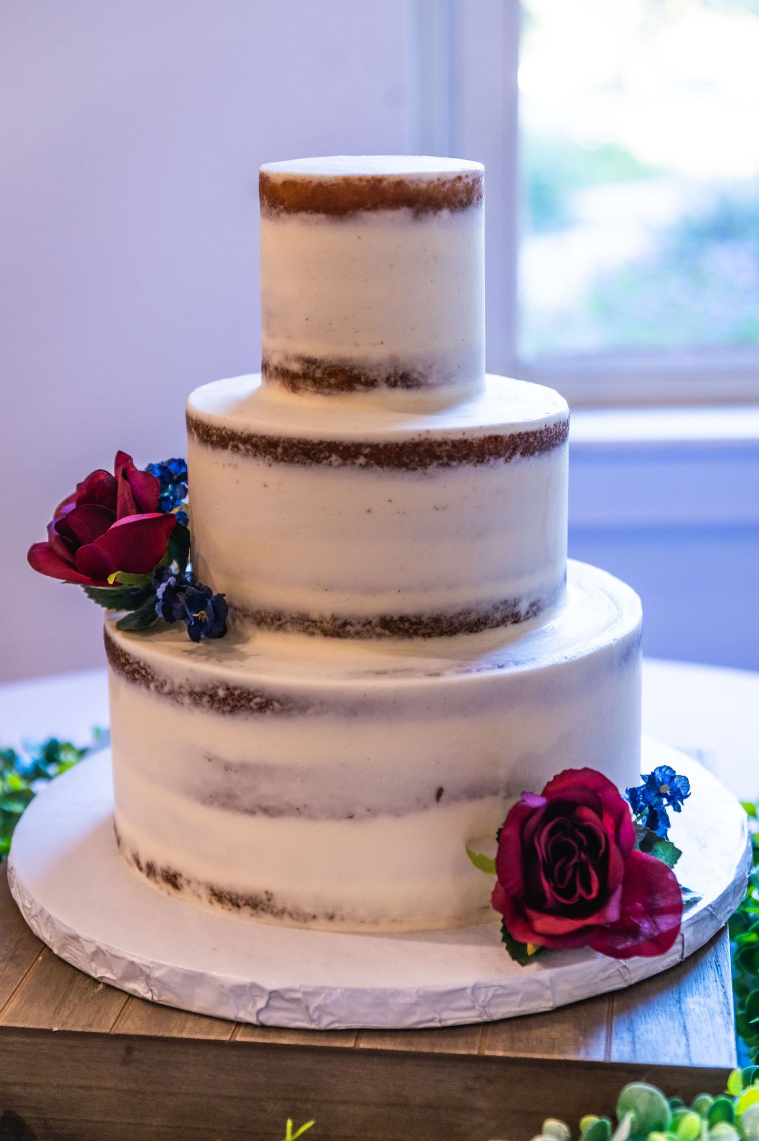 A three-tier semi-naked wedding cake decorated with burgundy roses and blue flowers.