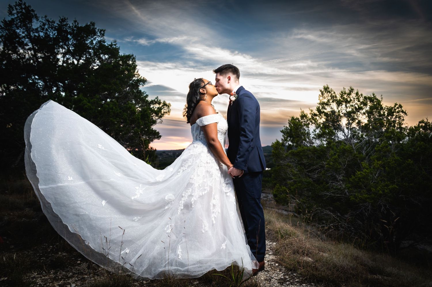 Dramatic sunset wedding photo with bride's dress flowing in the wind on a hilltop.