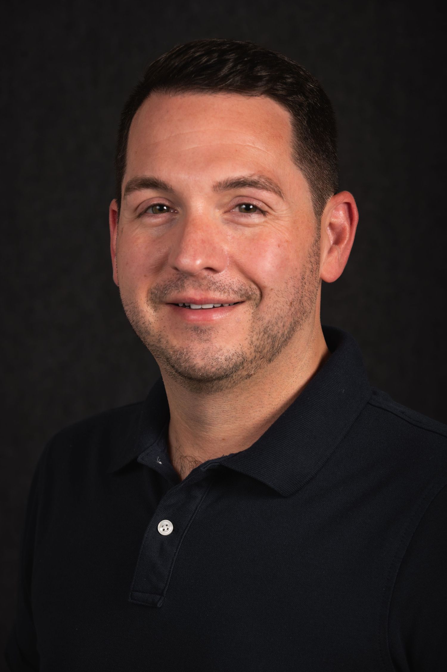 Professional headshot of a person in a navy polo shirt against dark background.