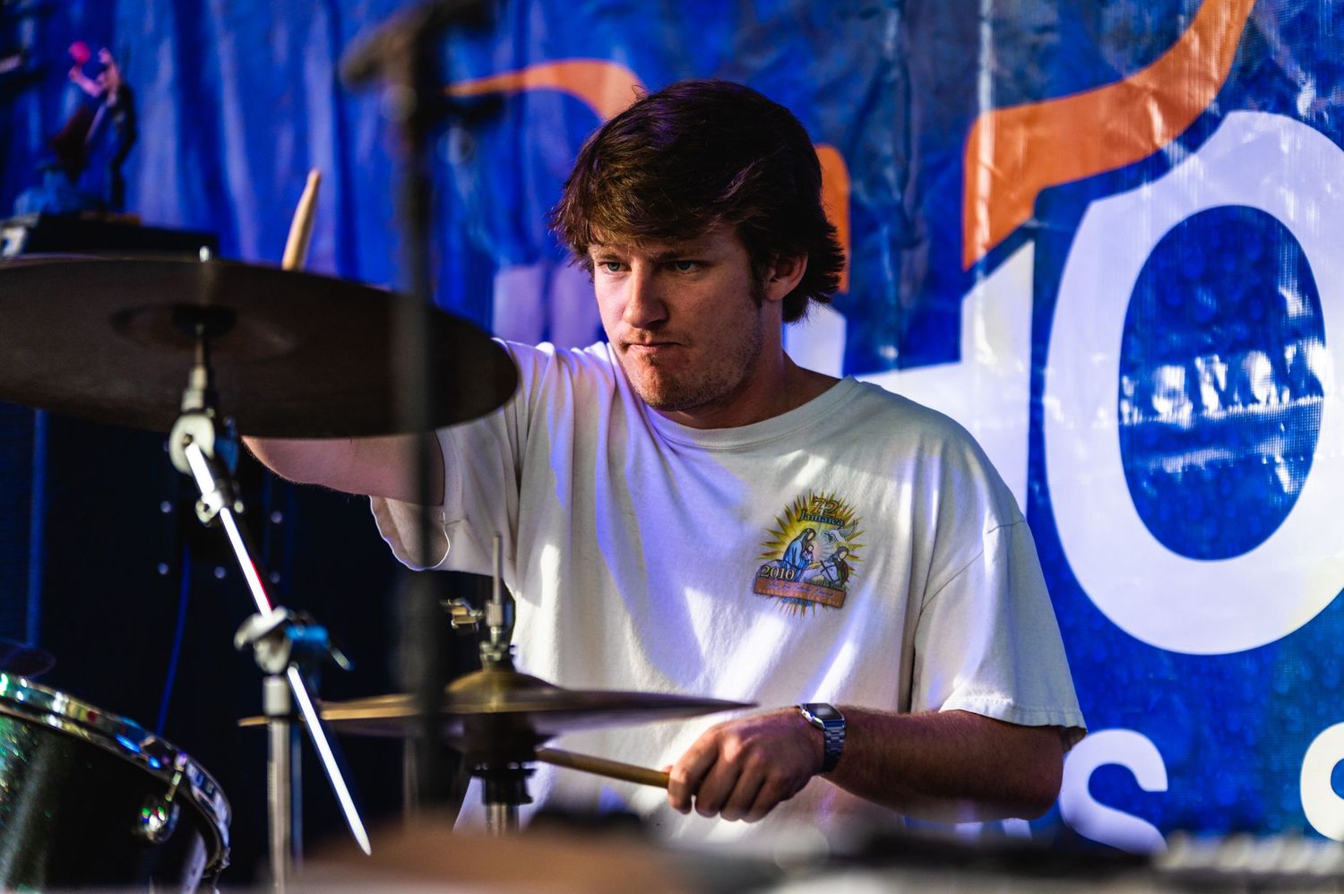 Drummer in white t-shirt performs behind drum kit during daytime outdoor show.