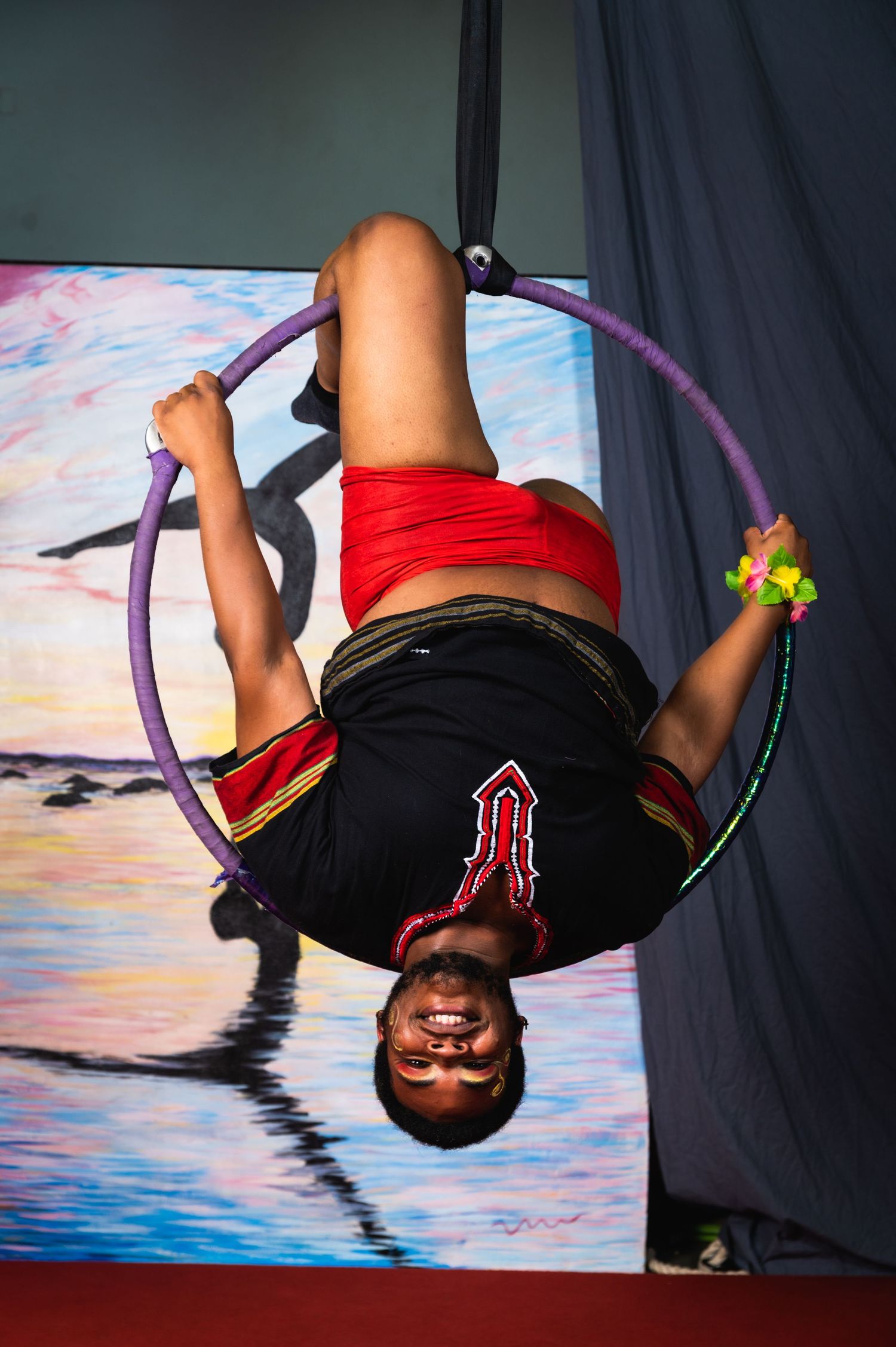 Acrobatic performer hangs upside down in a purple aerial hoop against sunset backdrop.