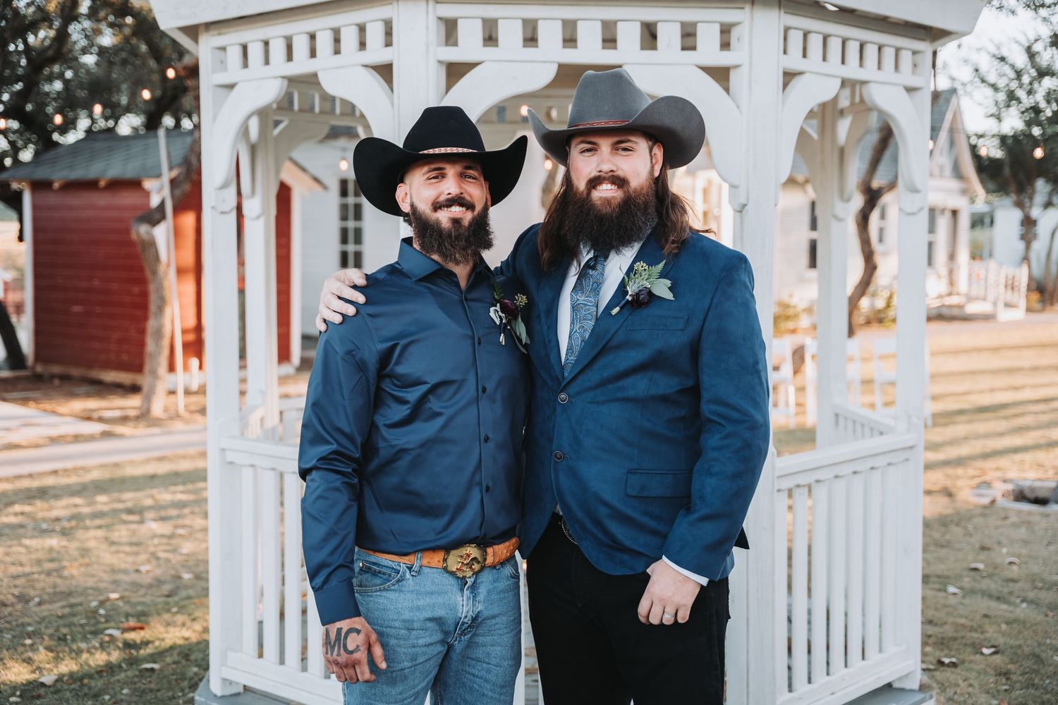 Two people in cowboy hats and blue suits pose together in front of a white decorative arch at an outdoor wedding.