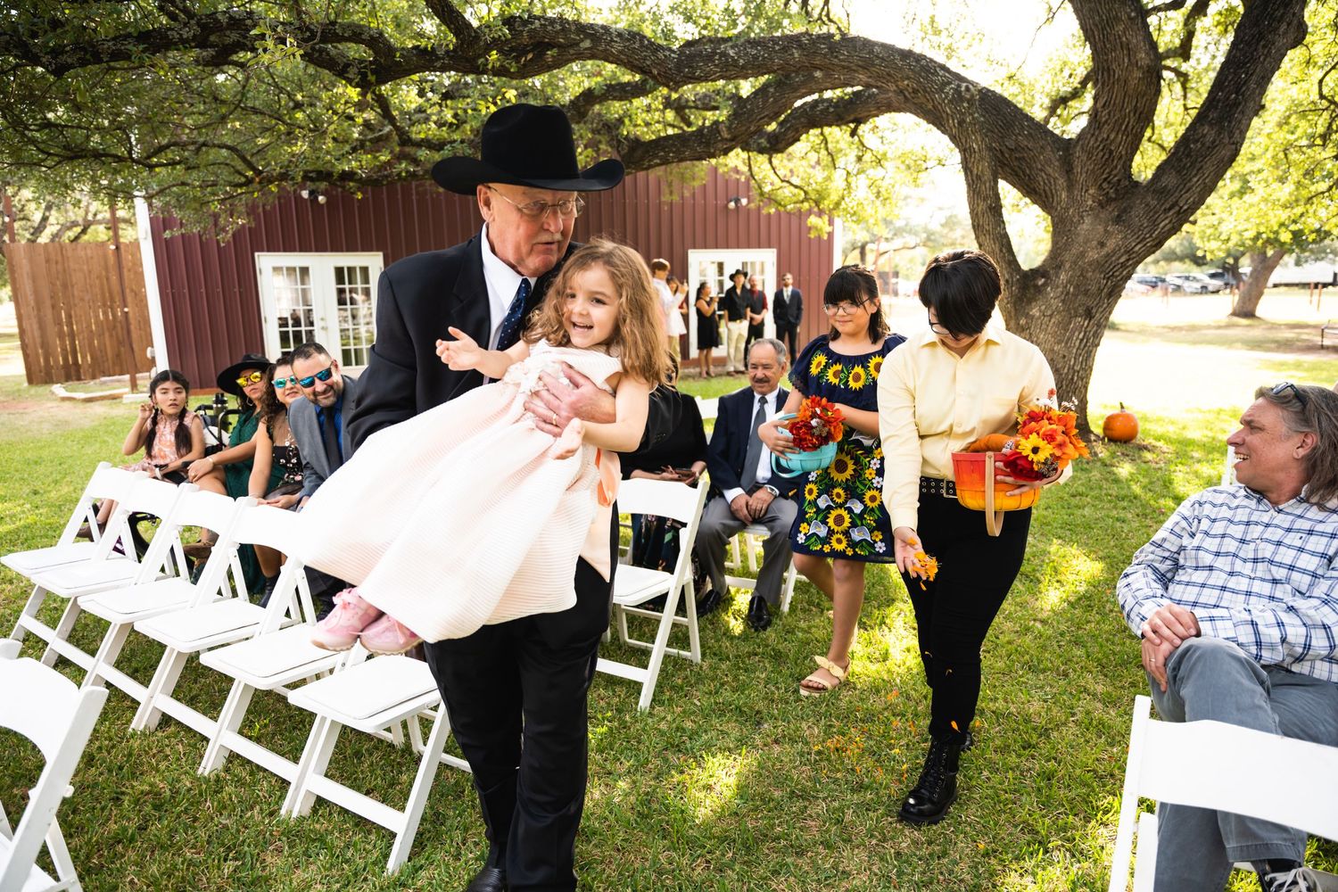 A tender moment during an outdoor ceremony with guests seated on white chairs beneath a shady tree.