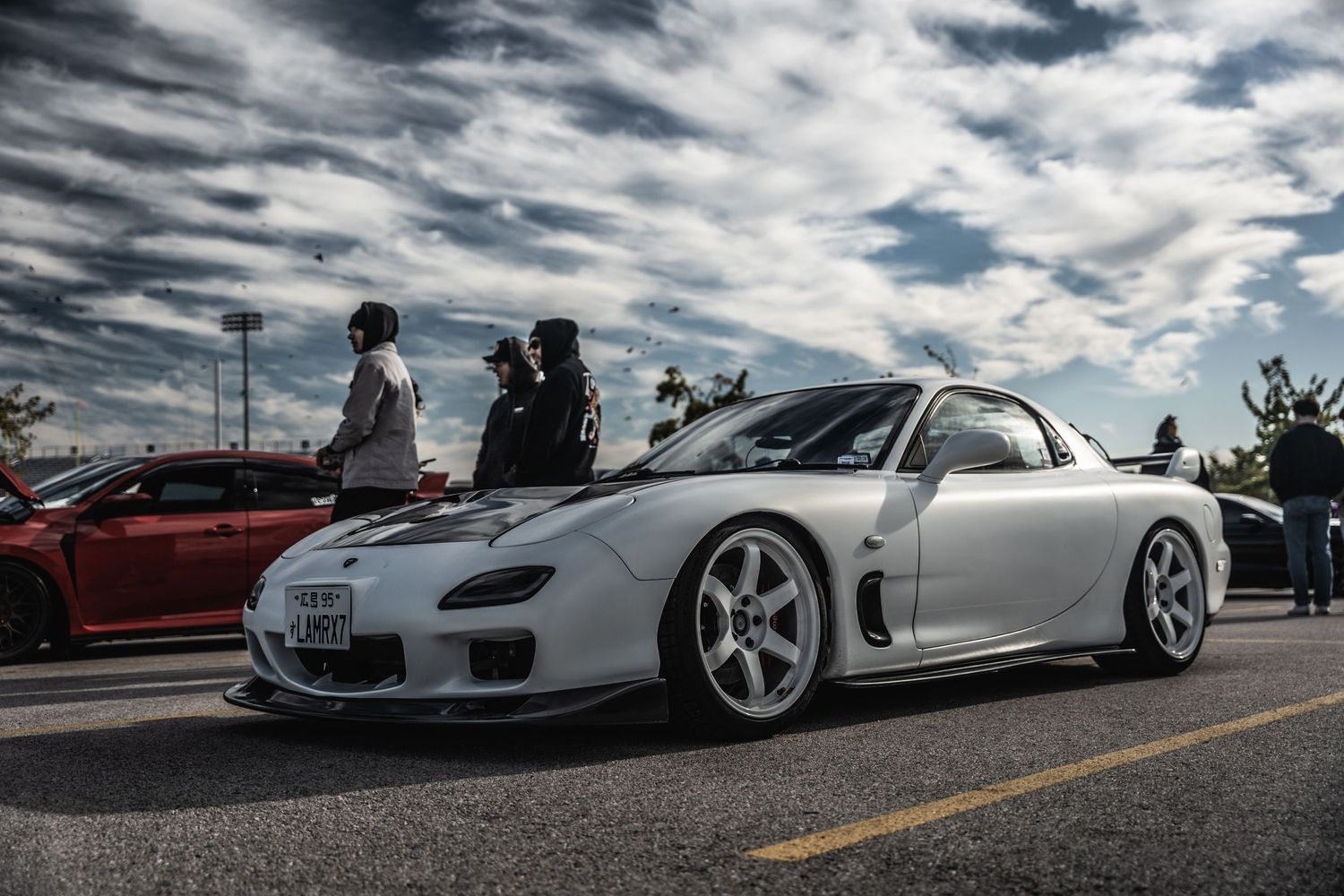 Modified white Mazda RX-7 with aftermarket wheels parked at a car show under dramatic cloudy skies.