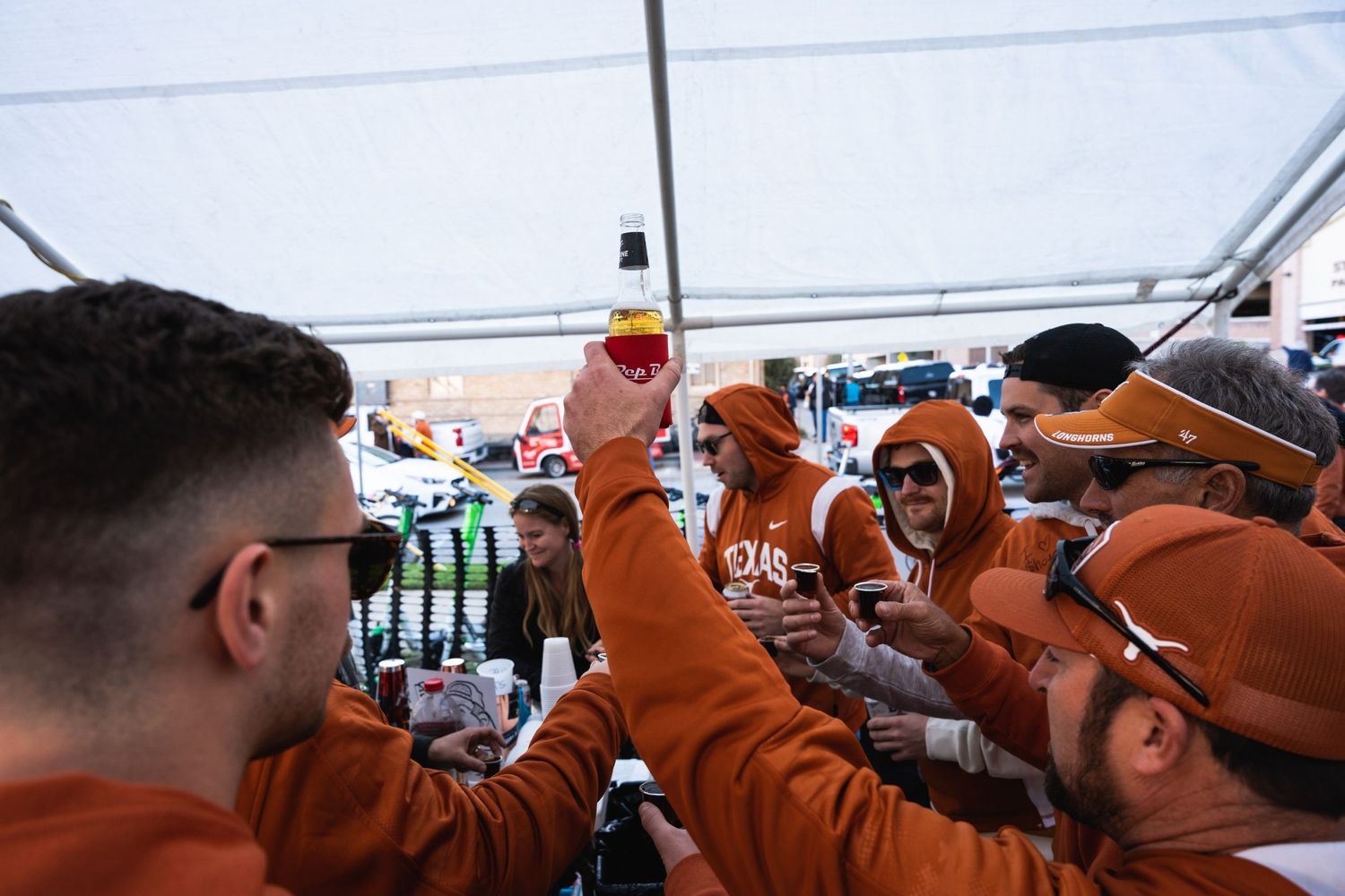Group wearing orange attire raises drinks in a celebratory toast under event tent.