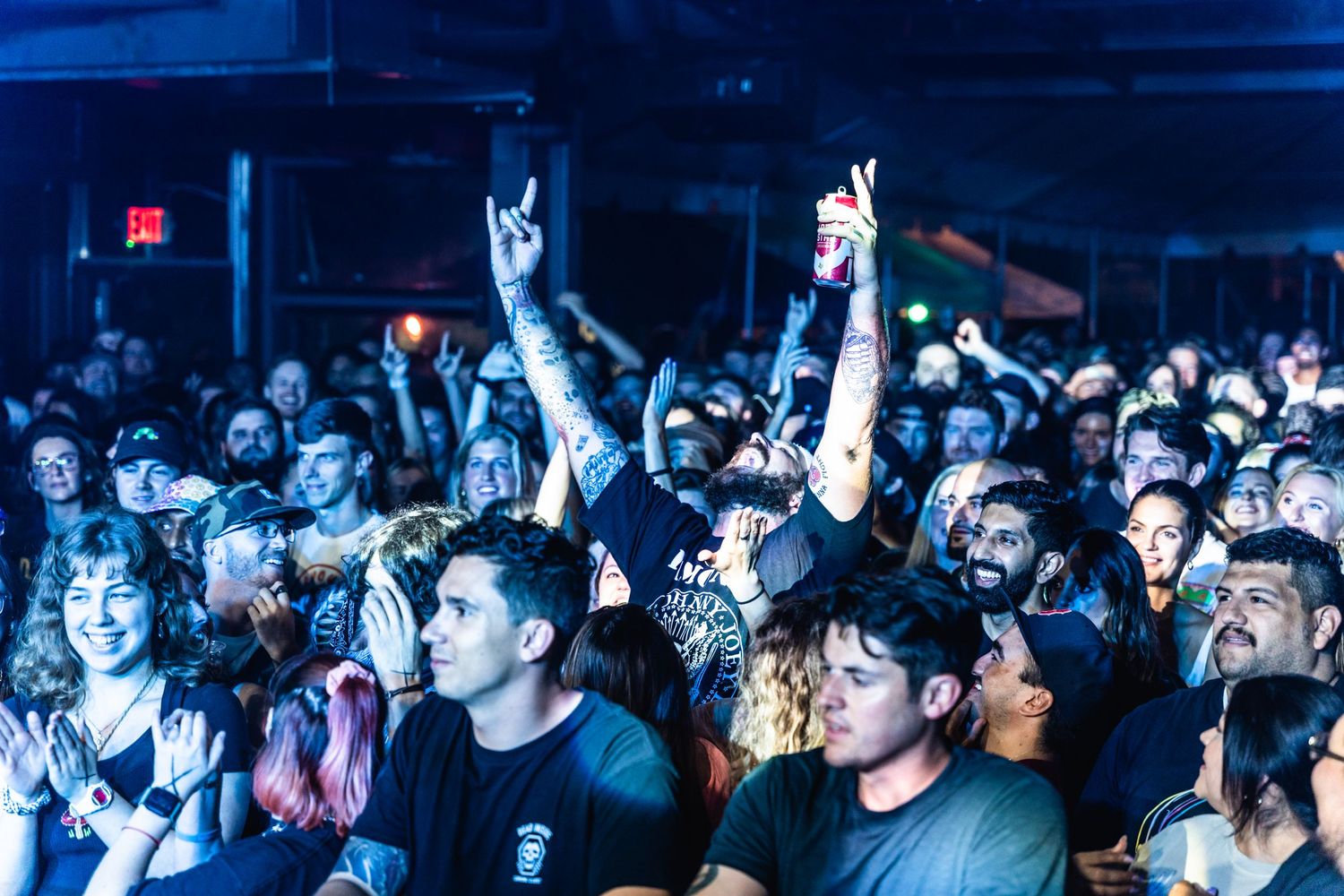 Energetic concert crowd raises devil horns hand gestures in a blue-lit venue.