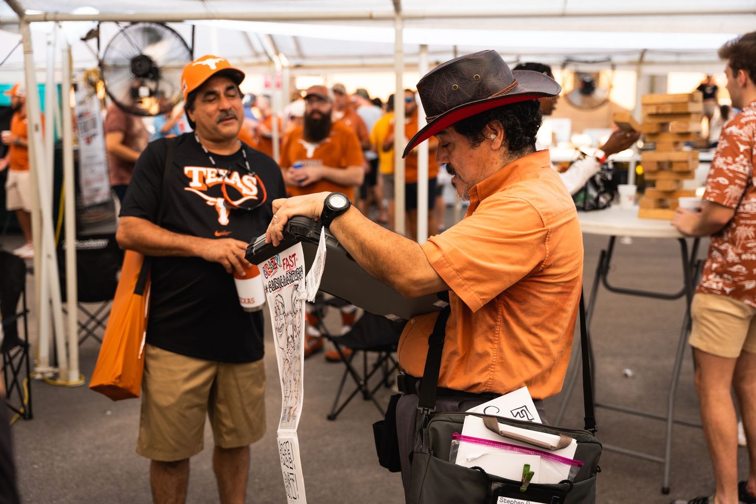 Two people interact at an outdoor event tent, with one wearing orange and another in a black t-shirt displaying artwork.