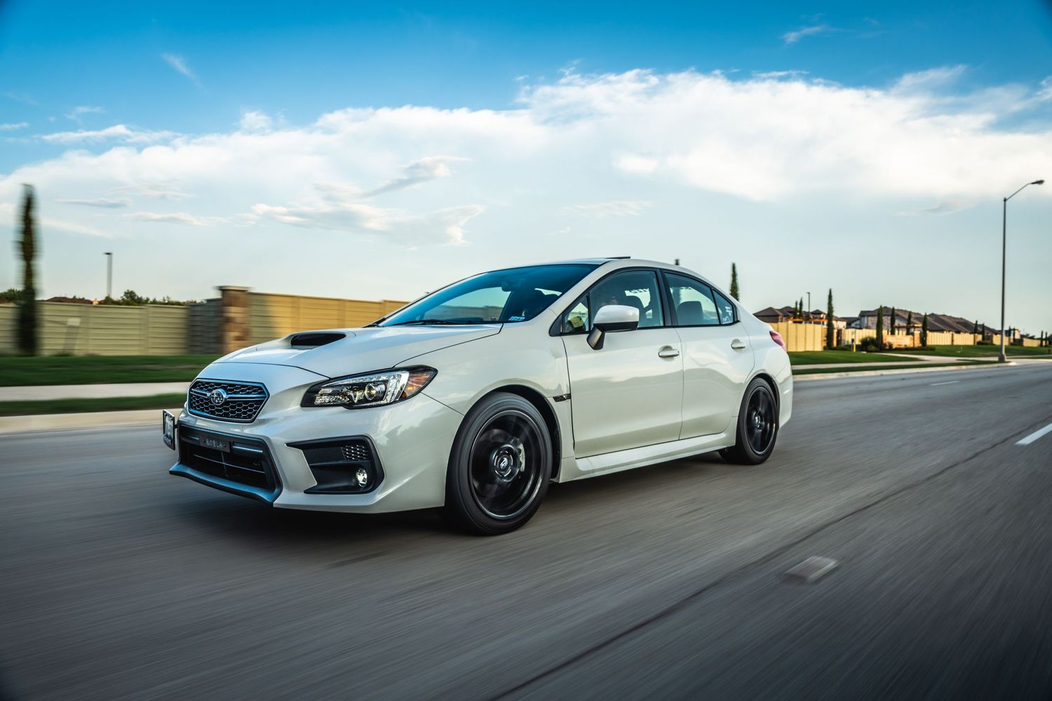 White Subaru WRX with black wheels photographed in motion on a suburban road.