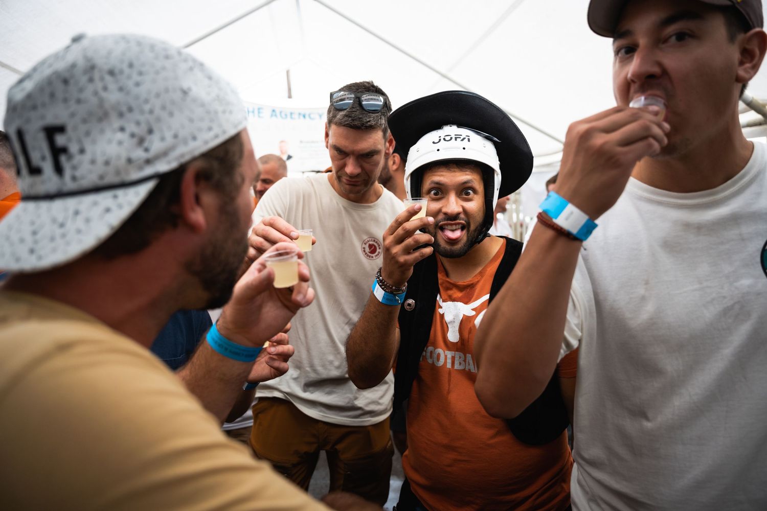 People socializing and taking shots at an outdoor drinking event under a tent.