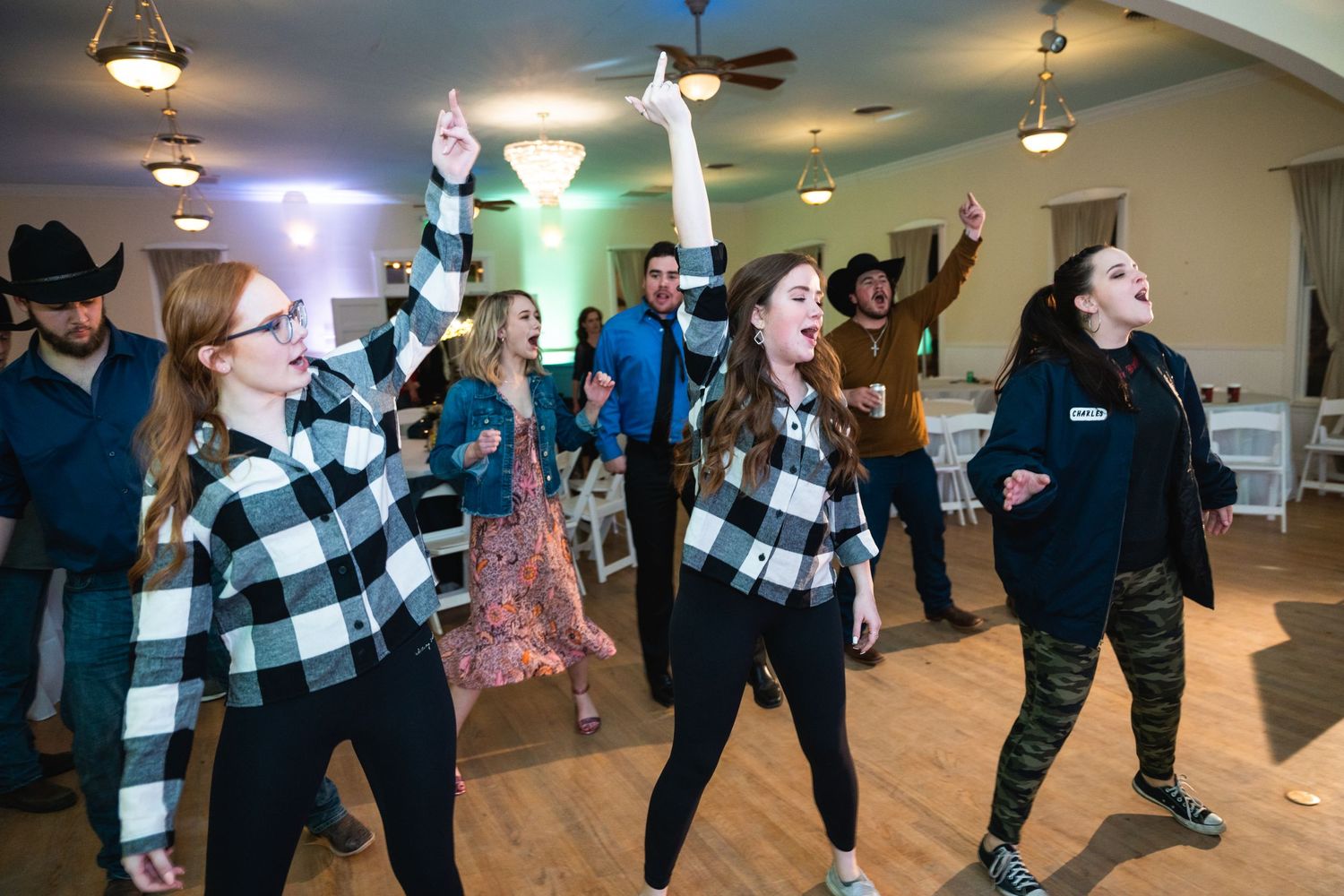 Wedding guests dancing in a line on wooden floor with plaid shirts and casual attire.