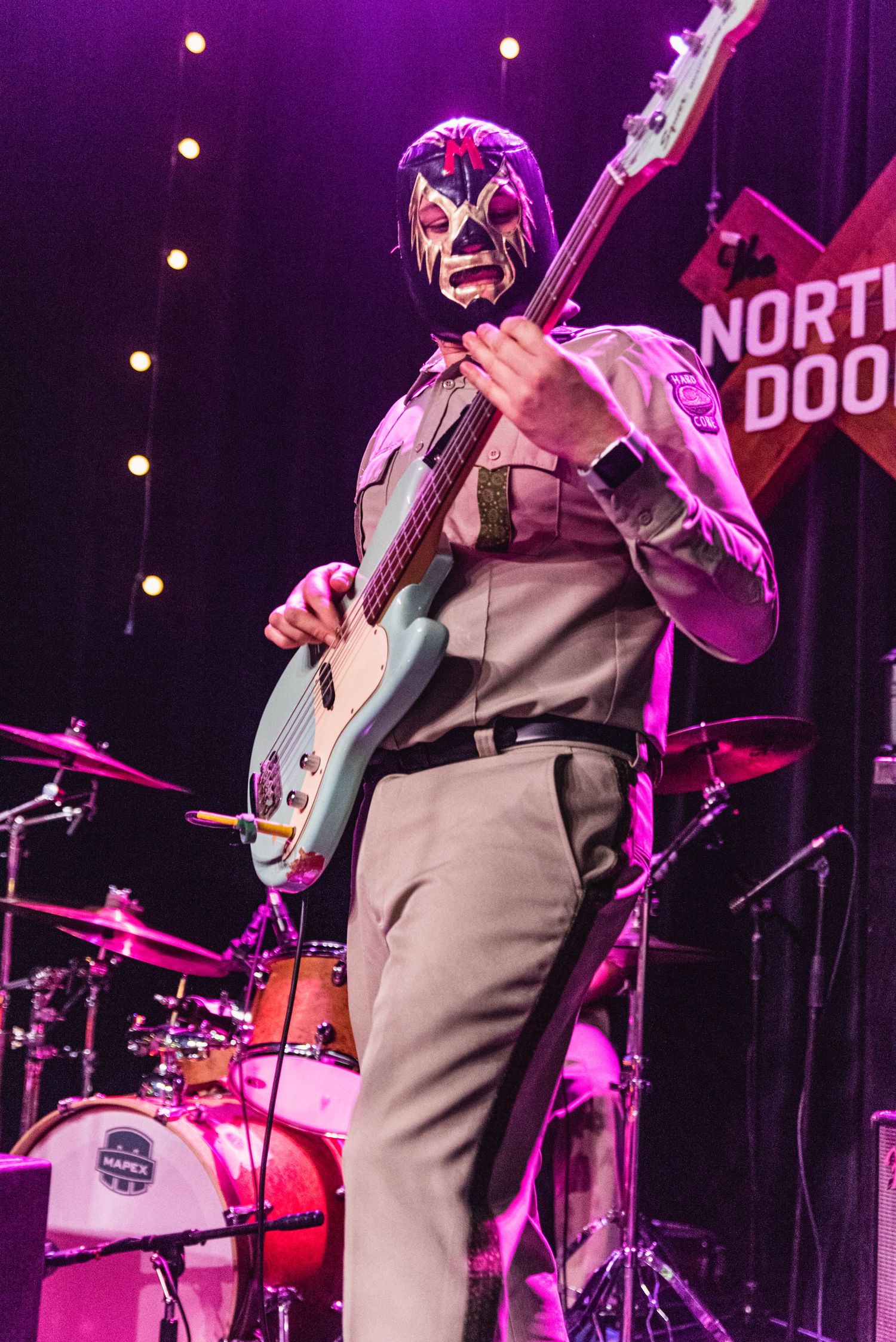 Bass guitarist wearing lucha libre mask performs on stage with purple lighting and drum kit visible.