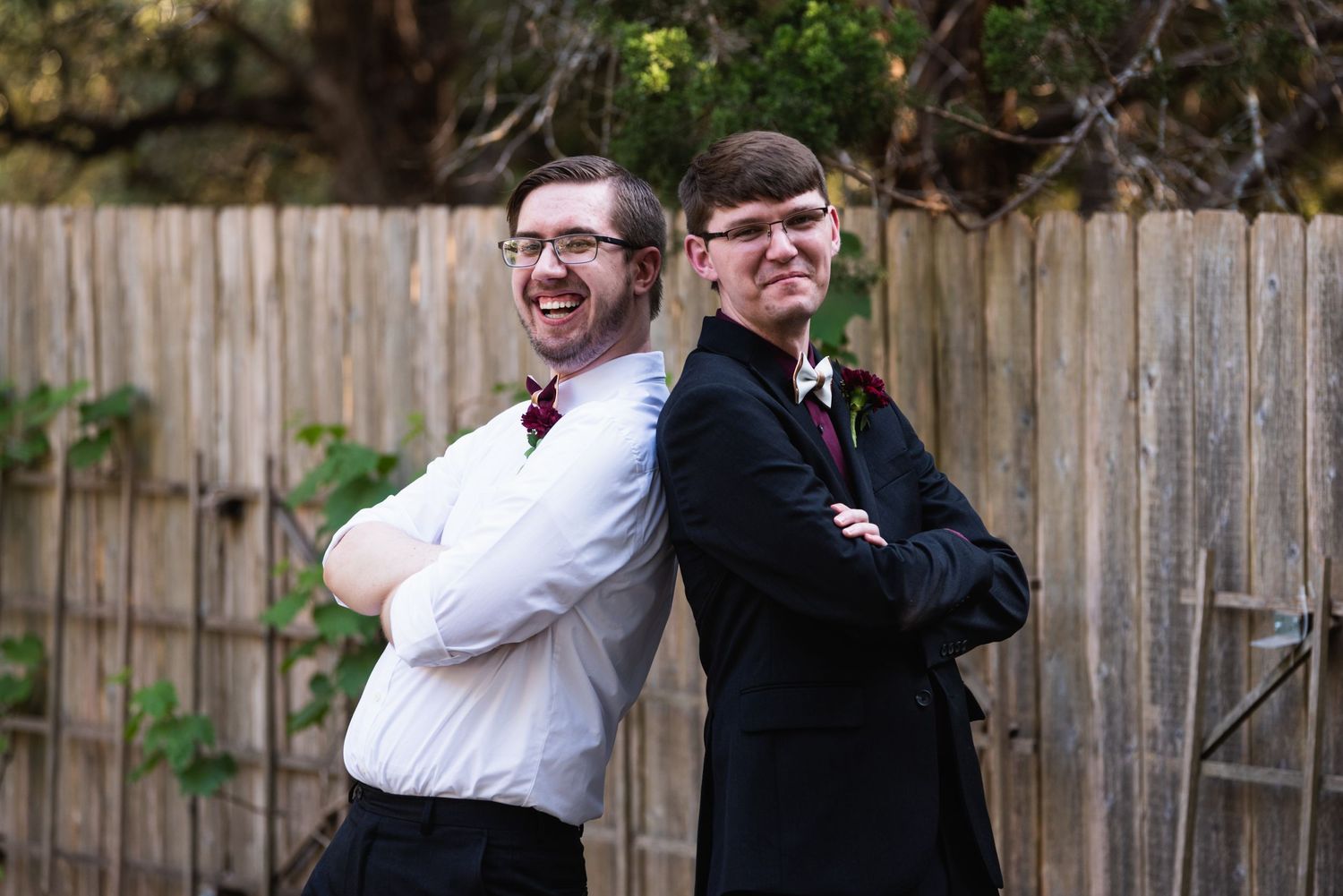 Two groomsmen pose back to back against a wooden fence wearing formal attire and bowties.