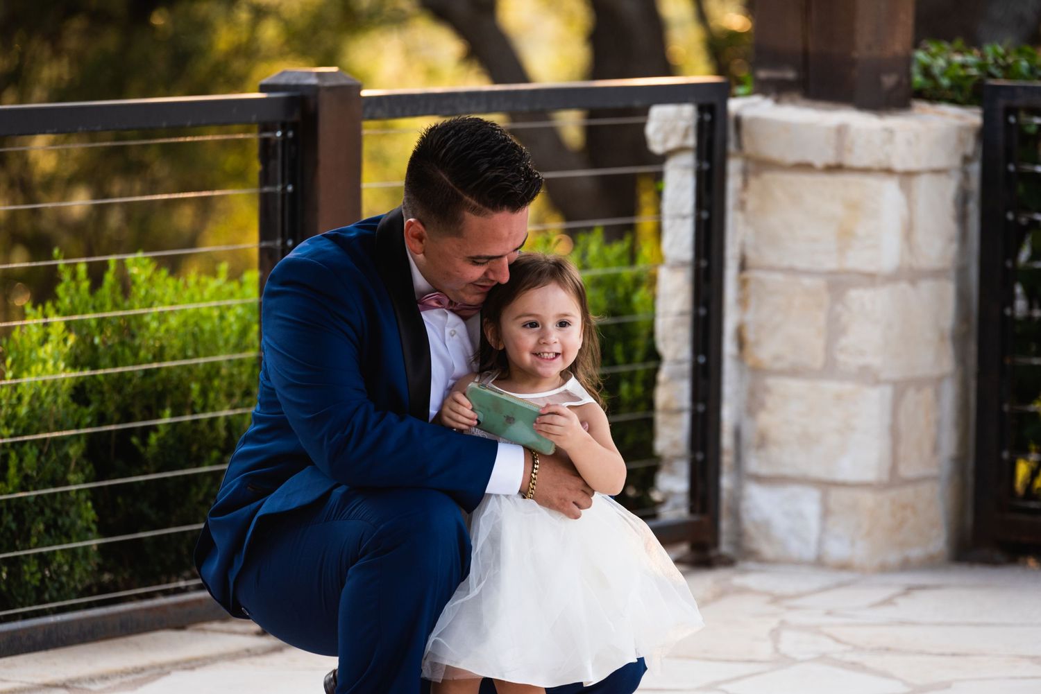 Person in blue suit embraces small child in white dress on outdoor patio.