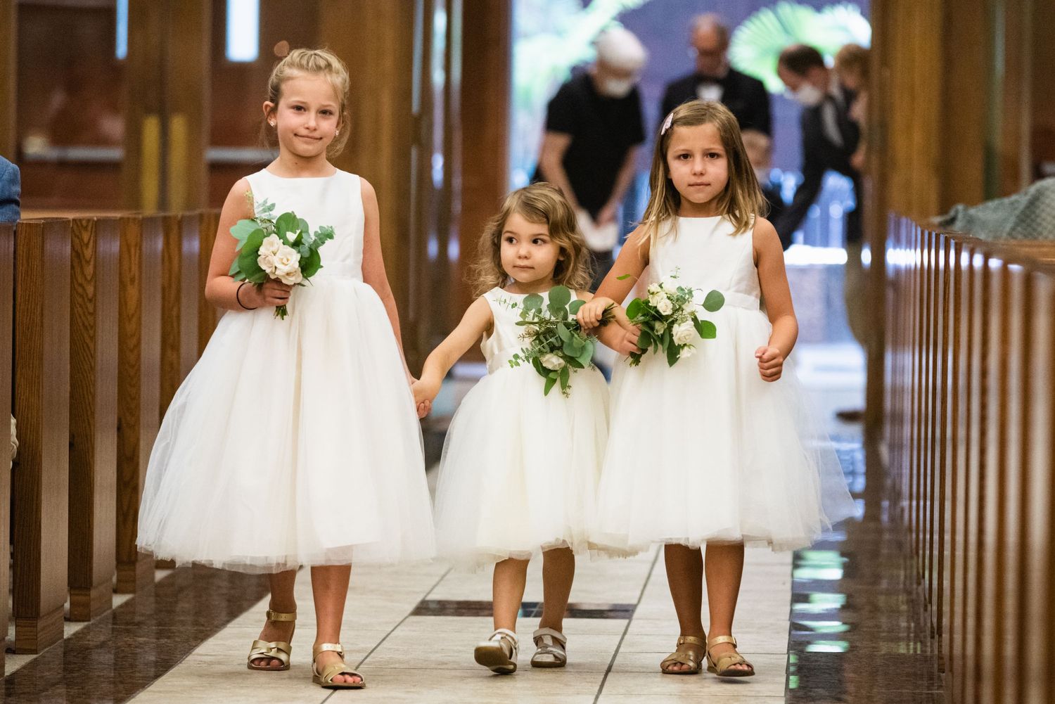 Three young flower girls in white dresses carrying bouquets walk down the church aisle.