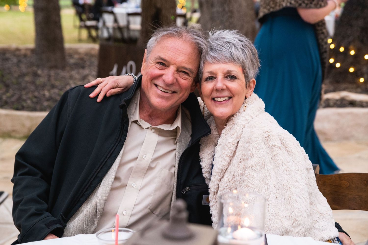 A happy mature couple smiles at an outdoor celebration with string lights visible in the background.