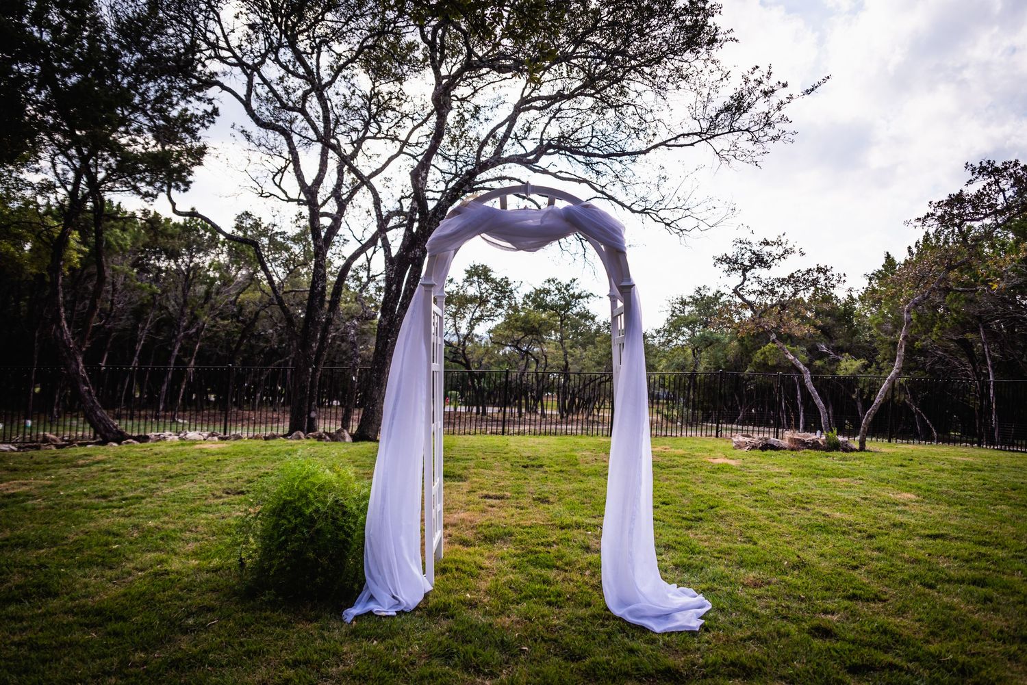 A simple white fabric wedding arch stands in a grassy field surrounded by trees.