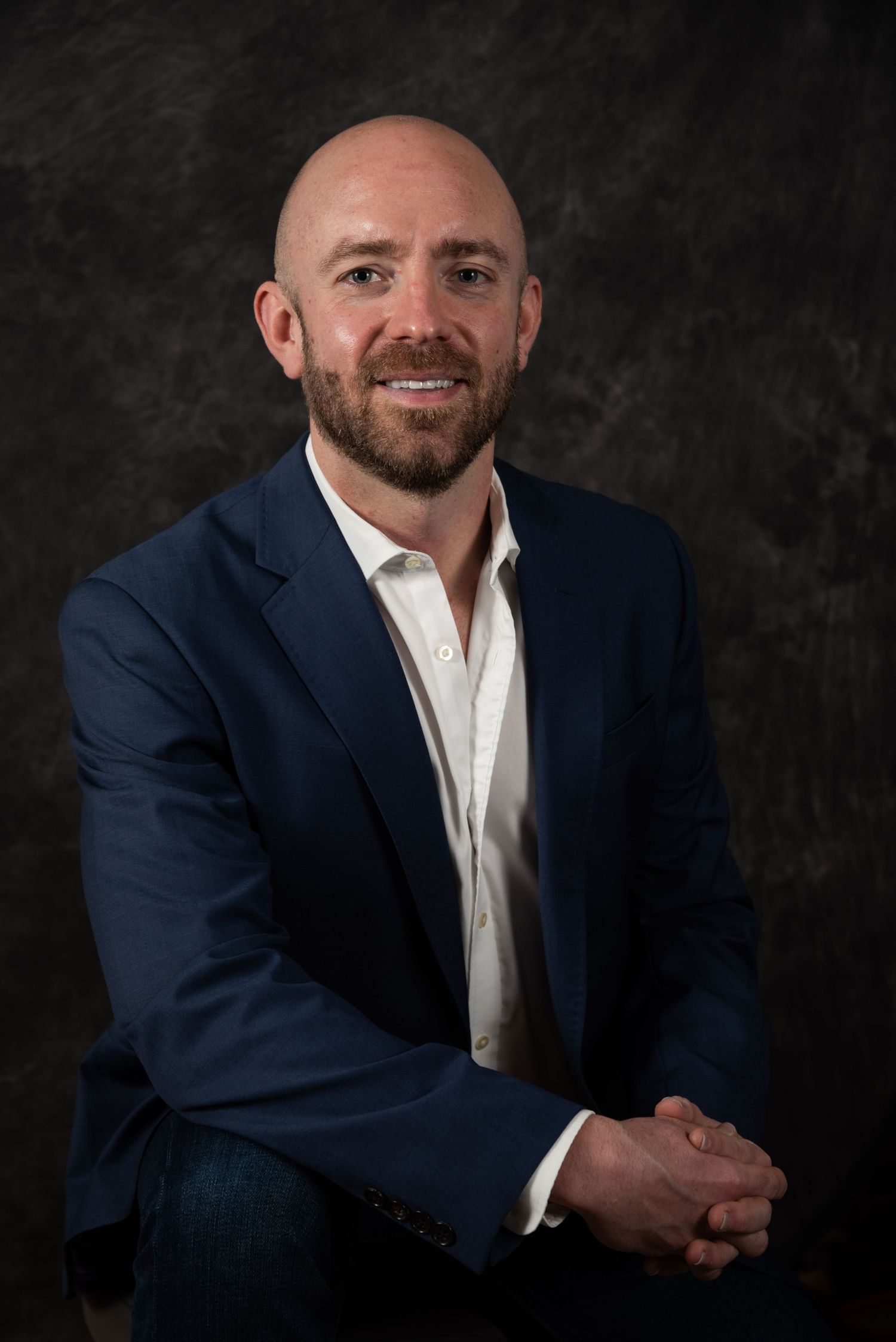 Professional seated portrait of person in navy blazer and white shirt against dark backdrop.