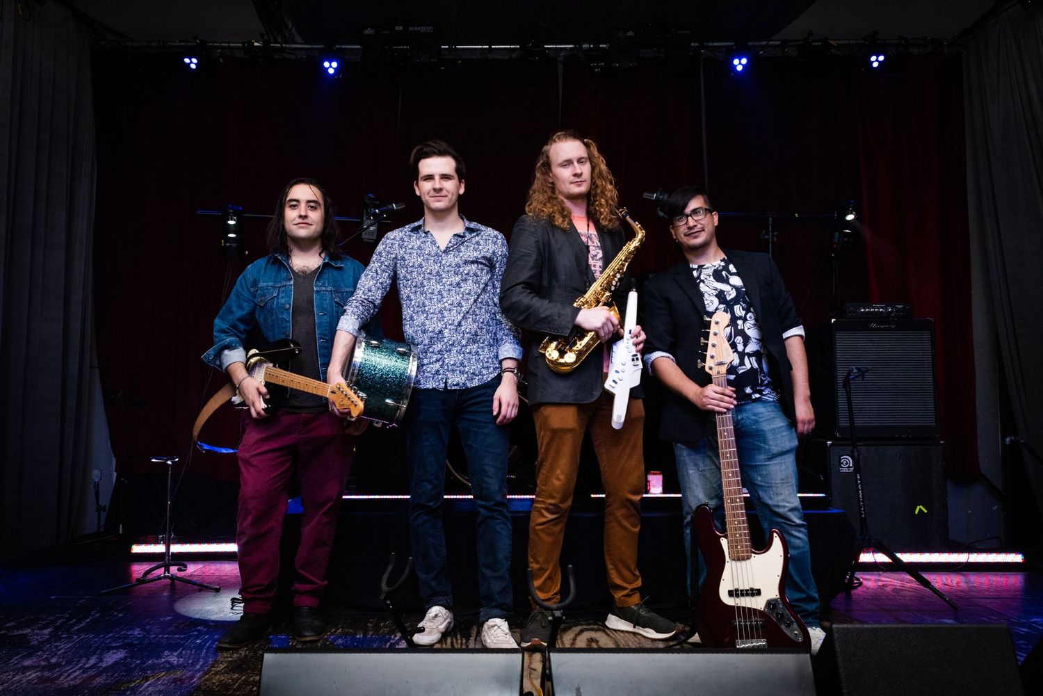 Four band members pose together on stage with their instruments in front of stage lighting.