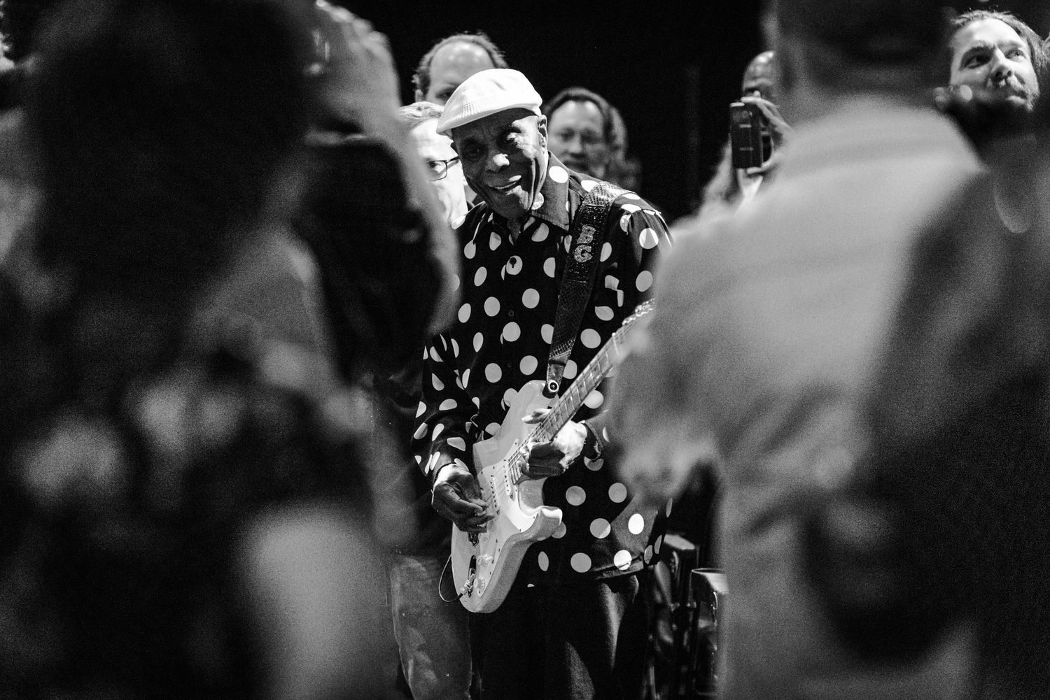Black and white concert photo showing performer in polka dot shirt through crowd of people.