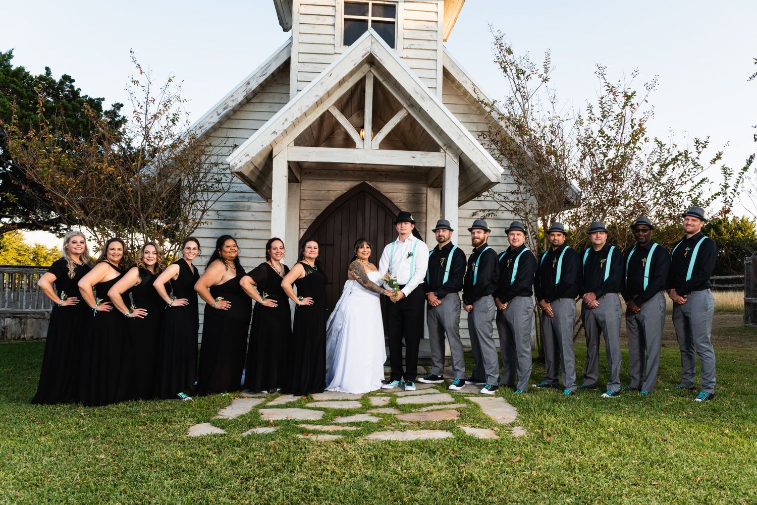 A wedding party poses in front of a white chapel with bridesmaids in black and groomsmen in gray.