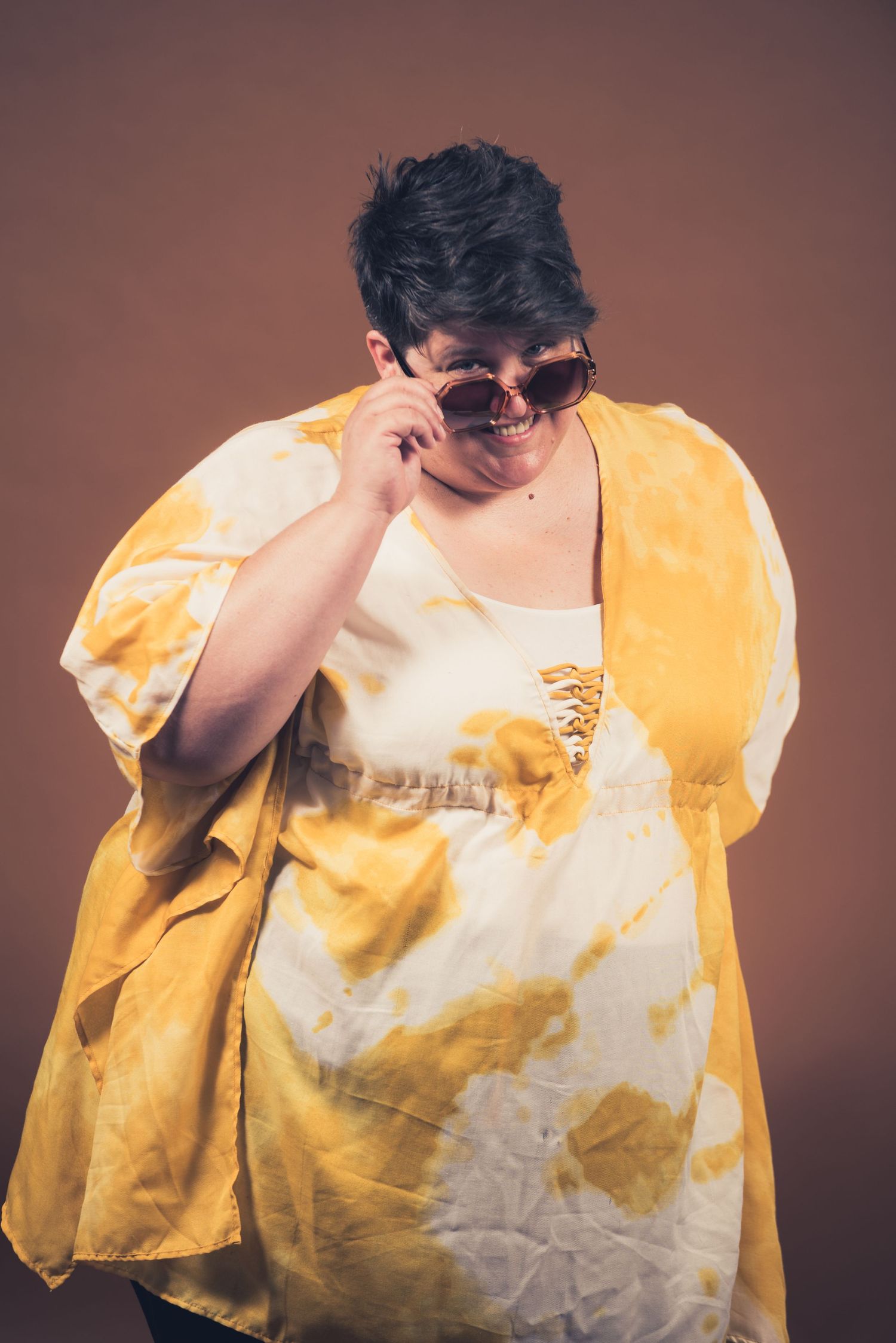 Person in yellow tie-dye outfit adjusting sunglasses against brown backdrop.