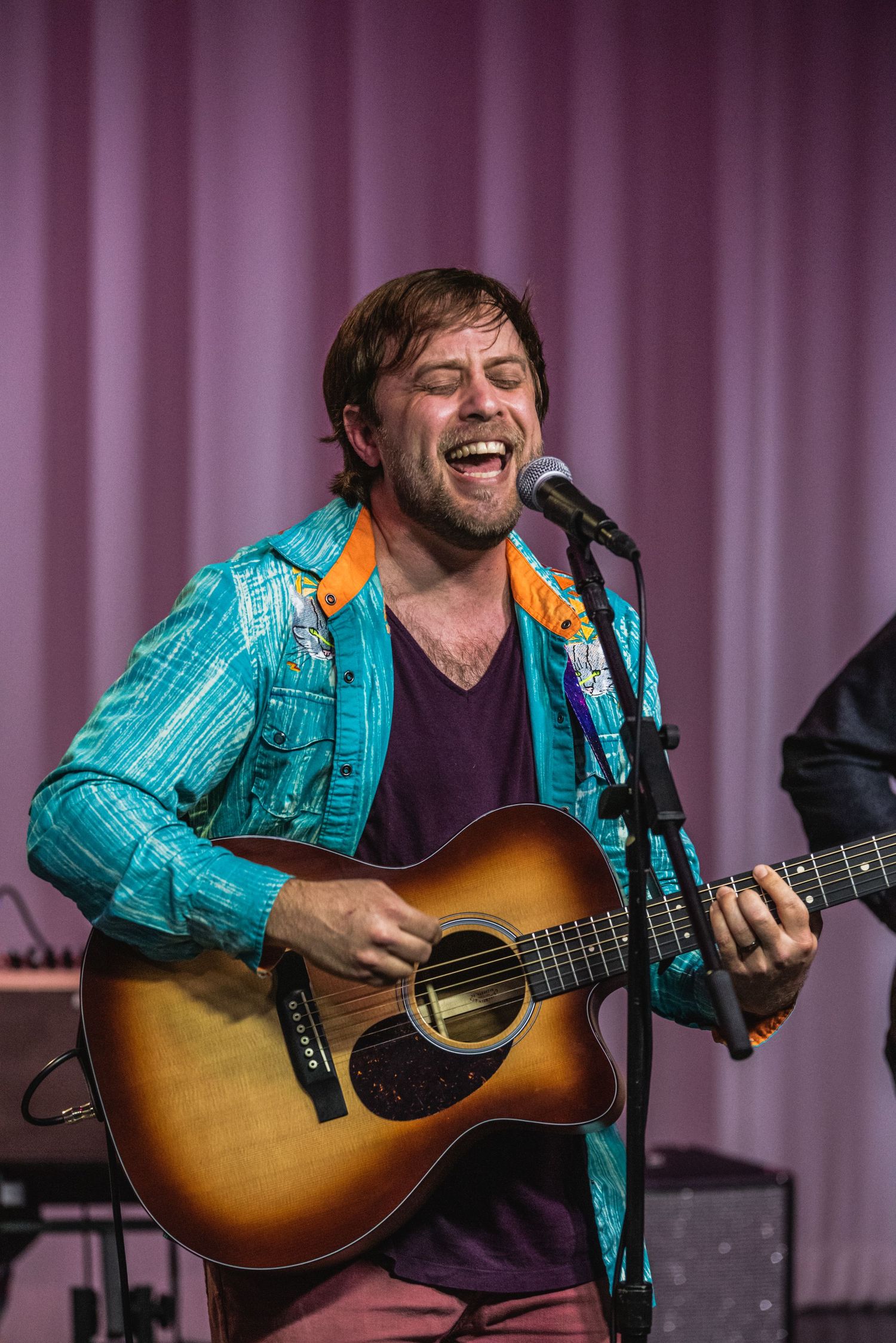 Musician in bright turquoise jacket sings passionately while playing acoustic guitar on stage.