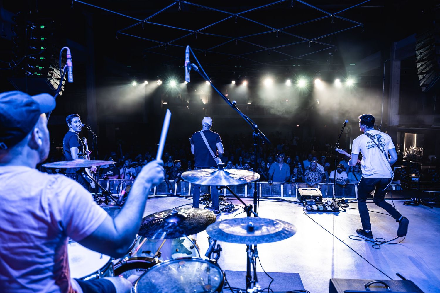 Drummer performing on stage with dramatic lighting and band members in background.