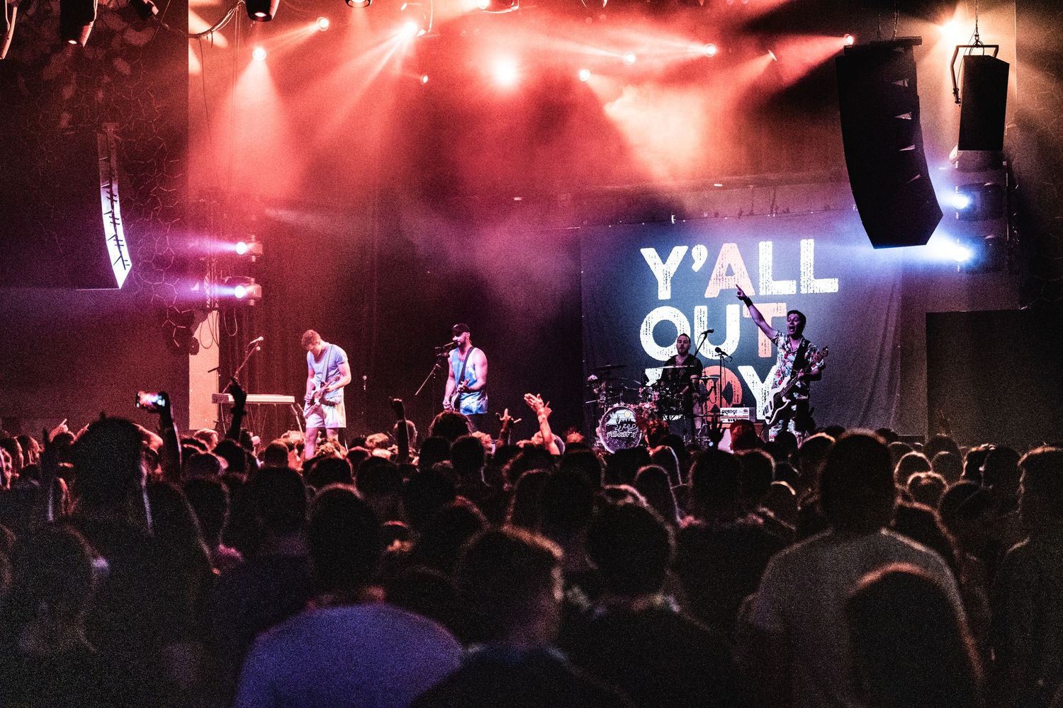 Concert crowd watches band perform under red lighting with 'Y'ALL OUT' sign visible on stage.