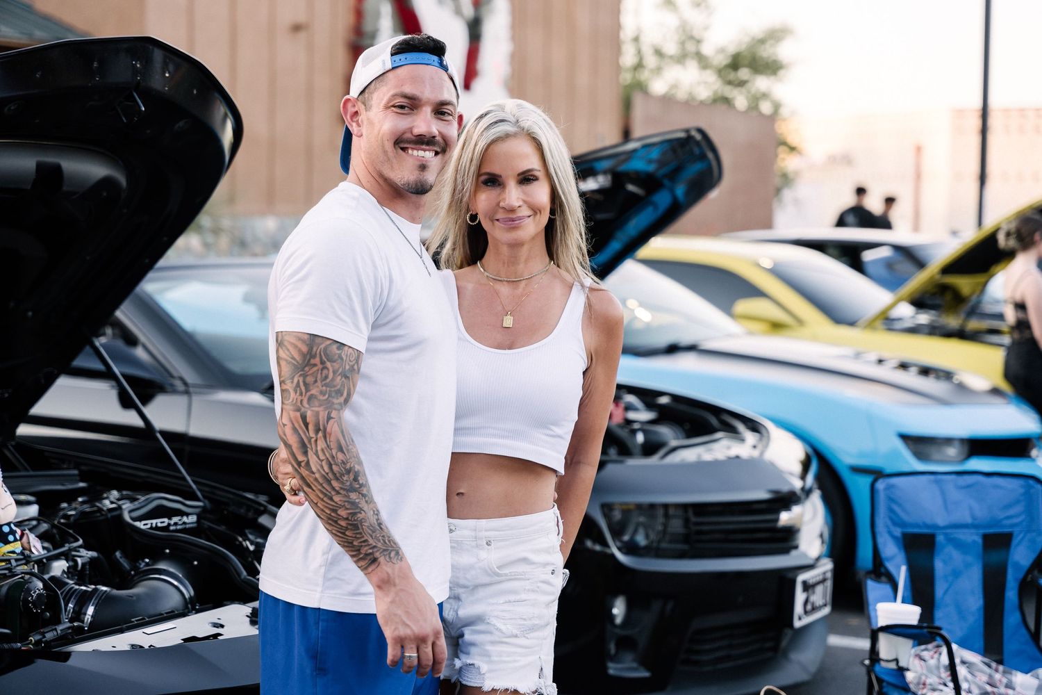 A couple in matching white outfits standing in front of cars with open hoods at an auto show.