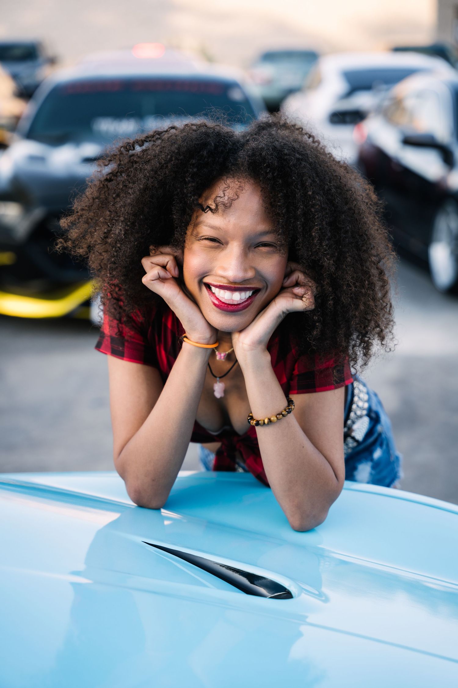 Person poses playfully on blue car hood at outdoor car meet.