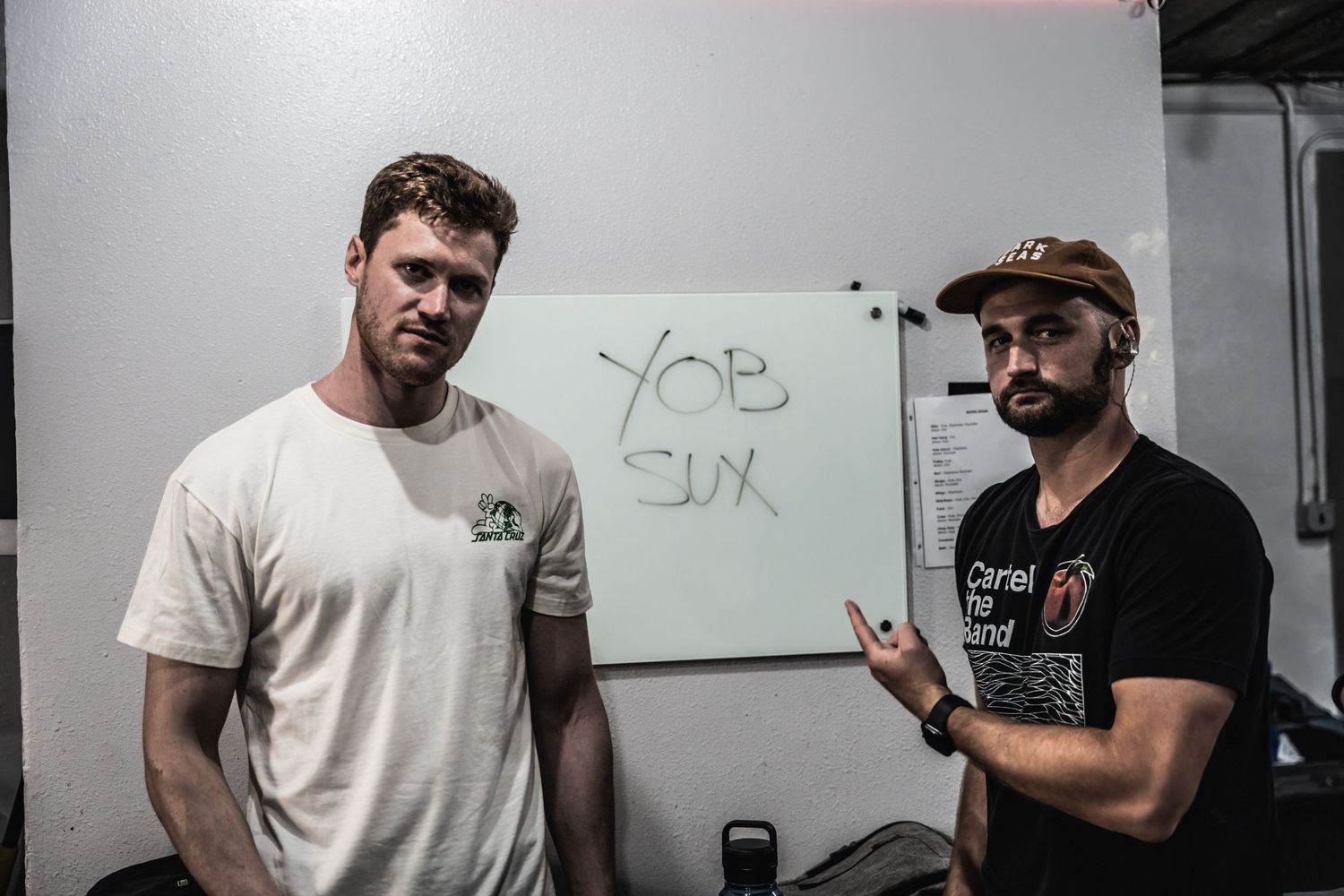 Two men standing in front of whiteboard in casual workout or gym setting.