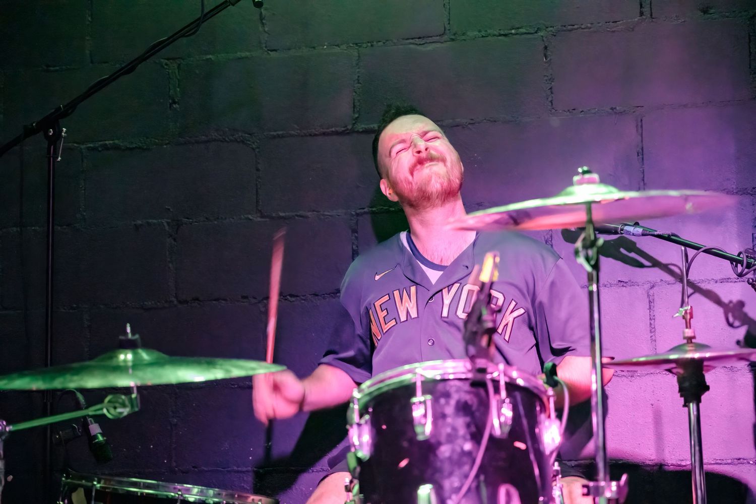 Drummer in New York shirt performs behind drum kit under purple and green stage lighting.