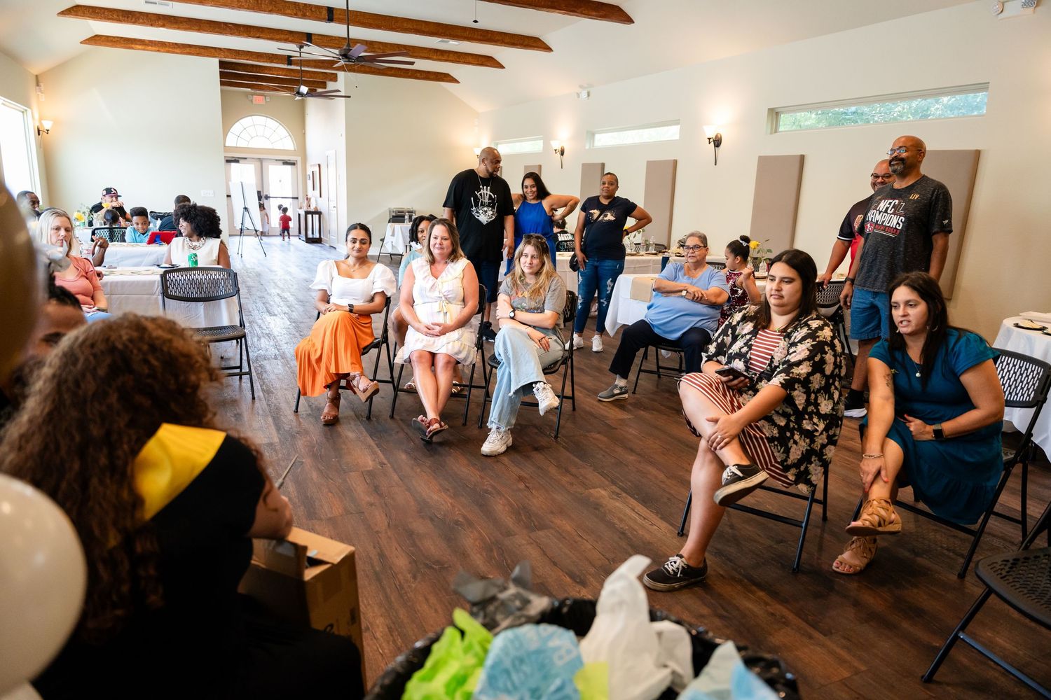 Wide view of a baby shower gathering in a room with wooden beams and guests seated in chairs.