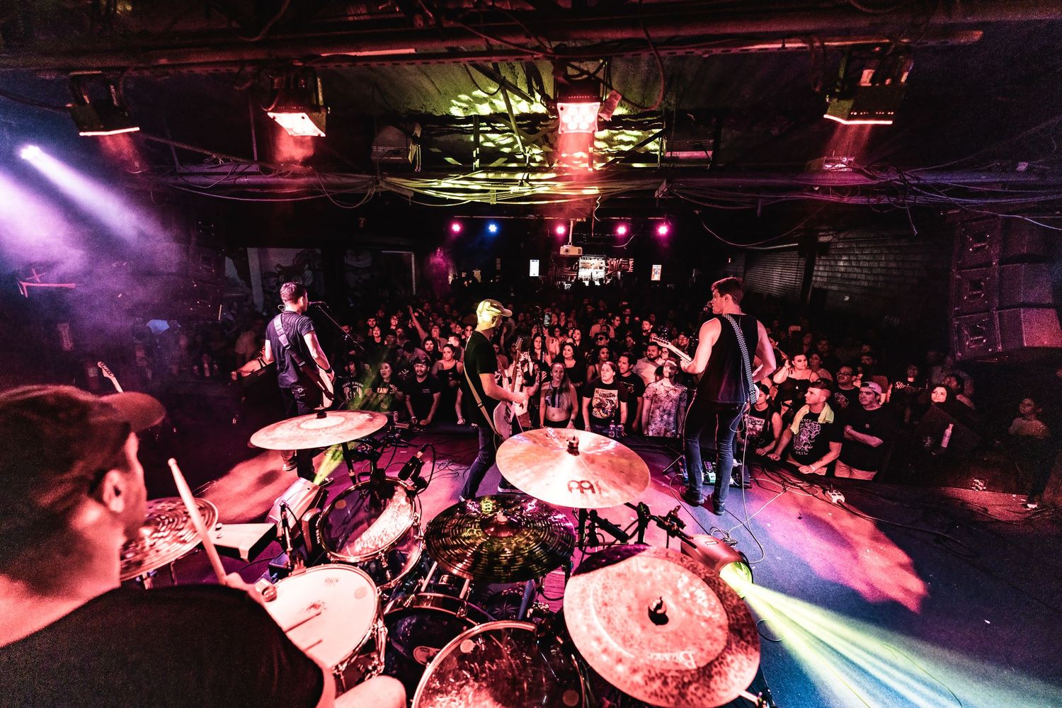 Drummer's perspective of a live concert with colorful stage lighting and packed audience.
